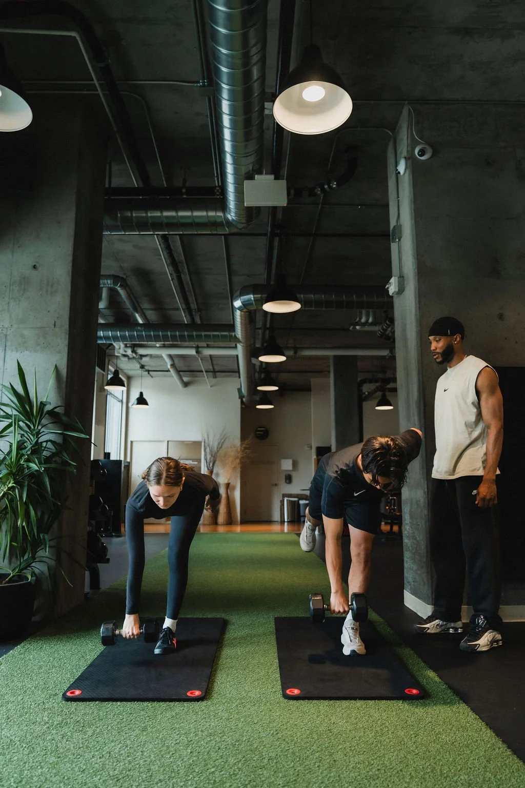 Two women exercising with dumbbells on workout mats, while a man watches indoors in a modern gym with exposed pipes and green turf flooring.