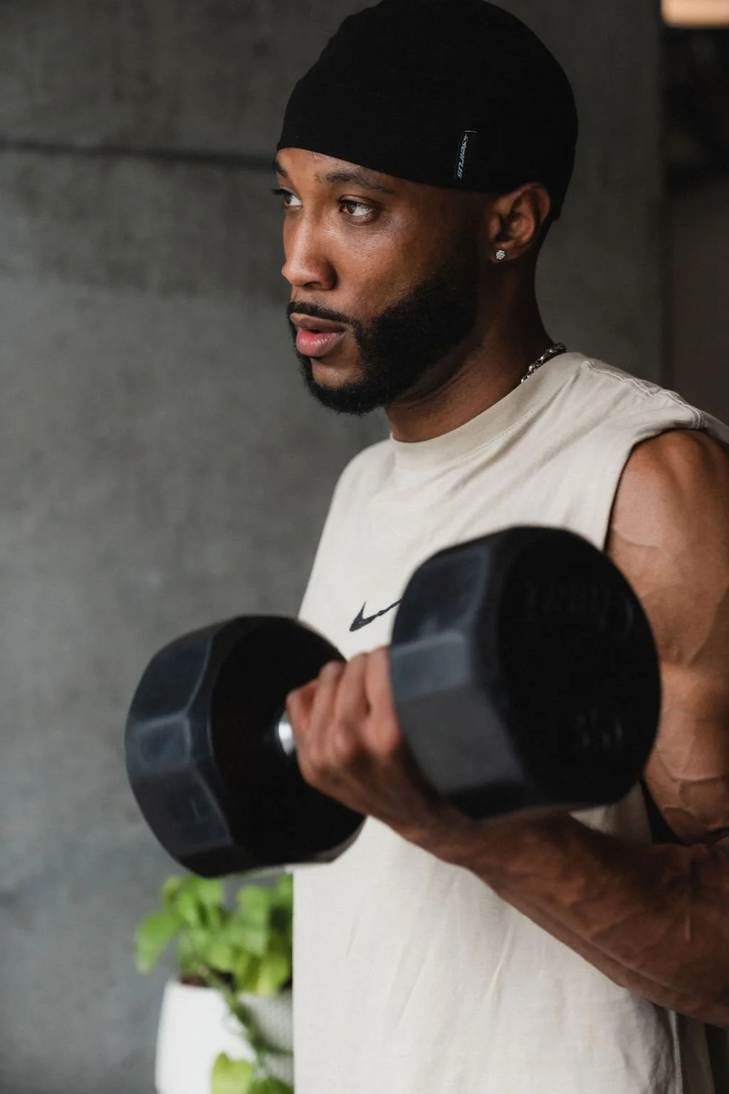 A man with a beard wearing a black head covering, sleeveless beige shirt, and earring, lifting a black dumbbell in a gym.