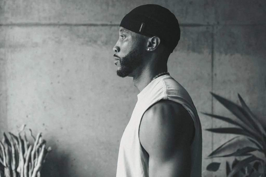 A black and white profile photo of a man wearing a sleeveless shirt, a watch, a chain necklace, and a beanie, standing against a concrete wall with plant leaves in the background.