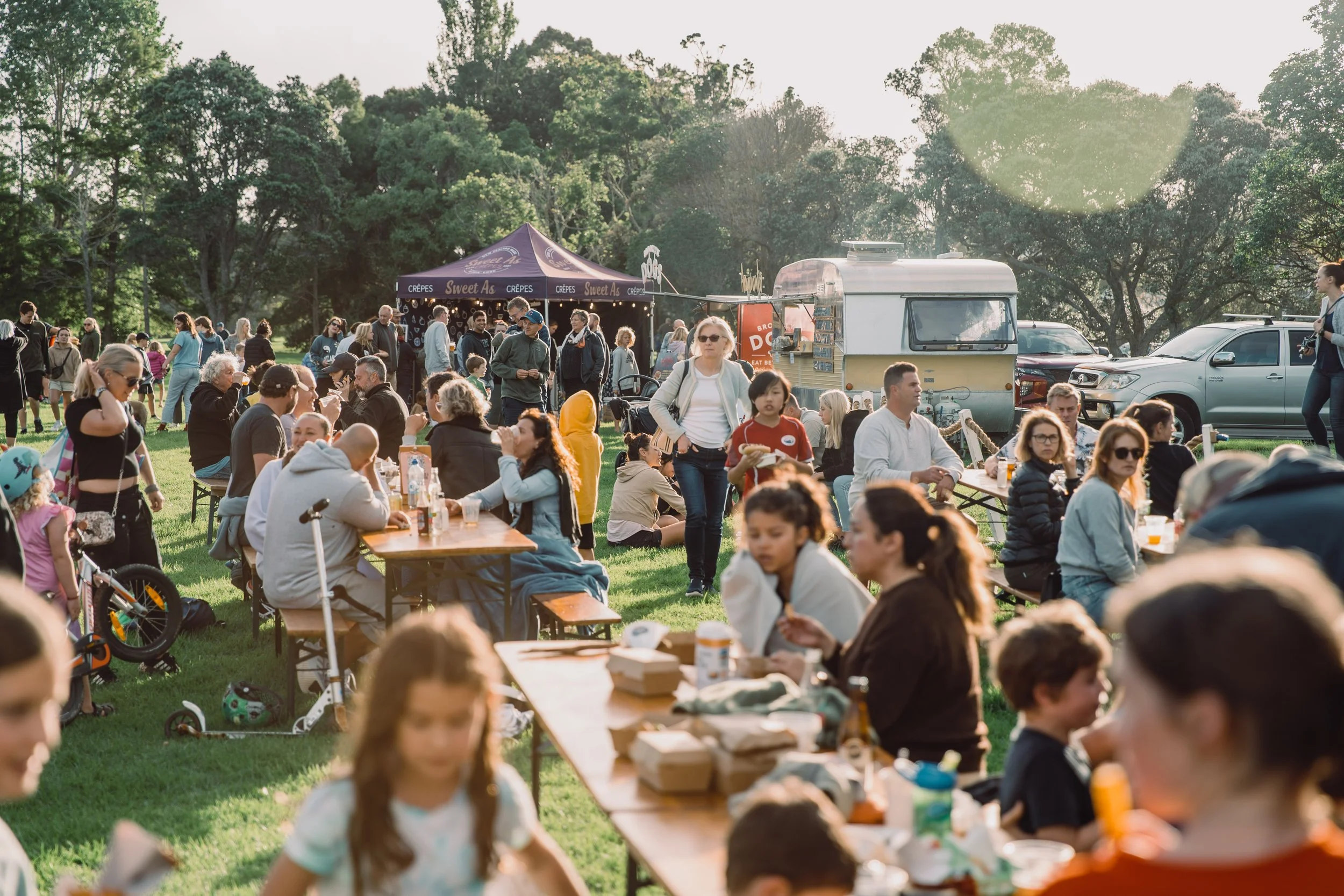 People gathered outdoors in a park with food stalls, tables, and cars, enjoying a sunny day.