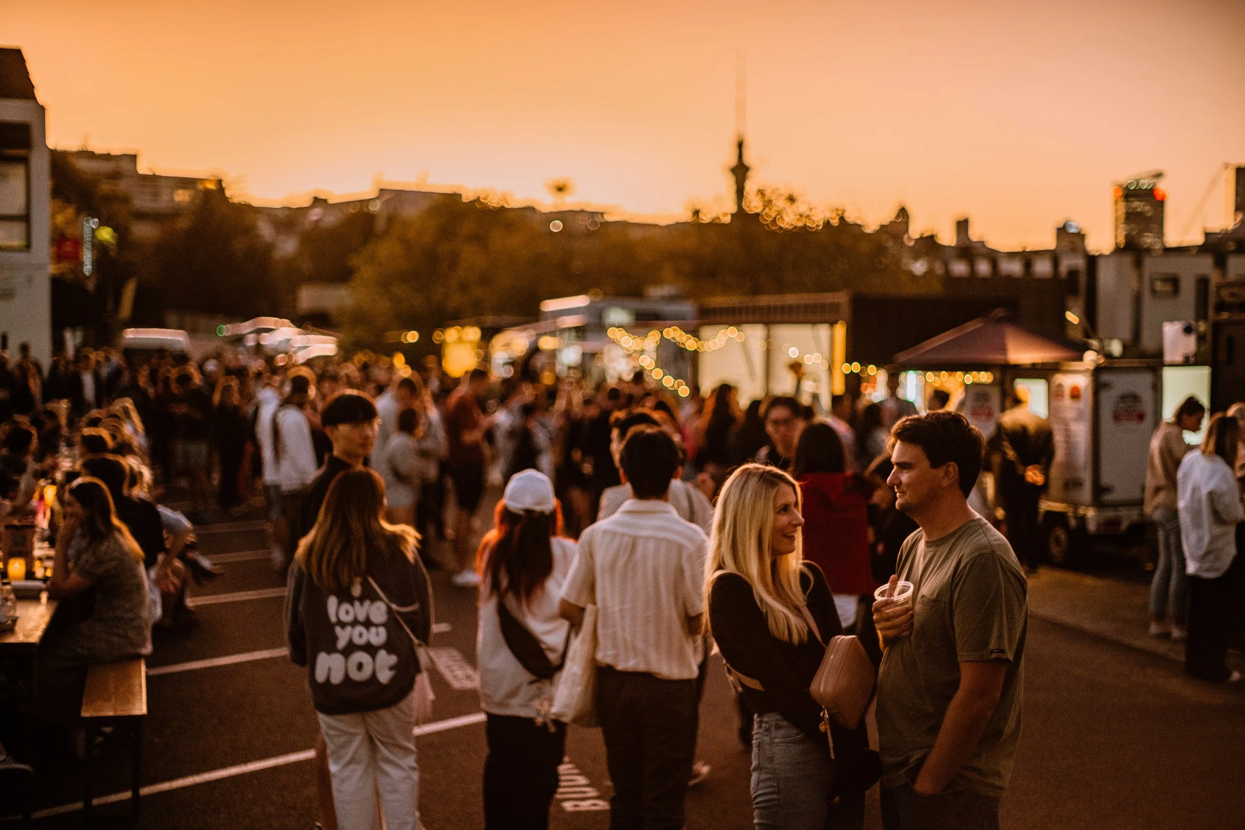 Crowd of people at an outdoor event during sunset with city skyline in the background.