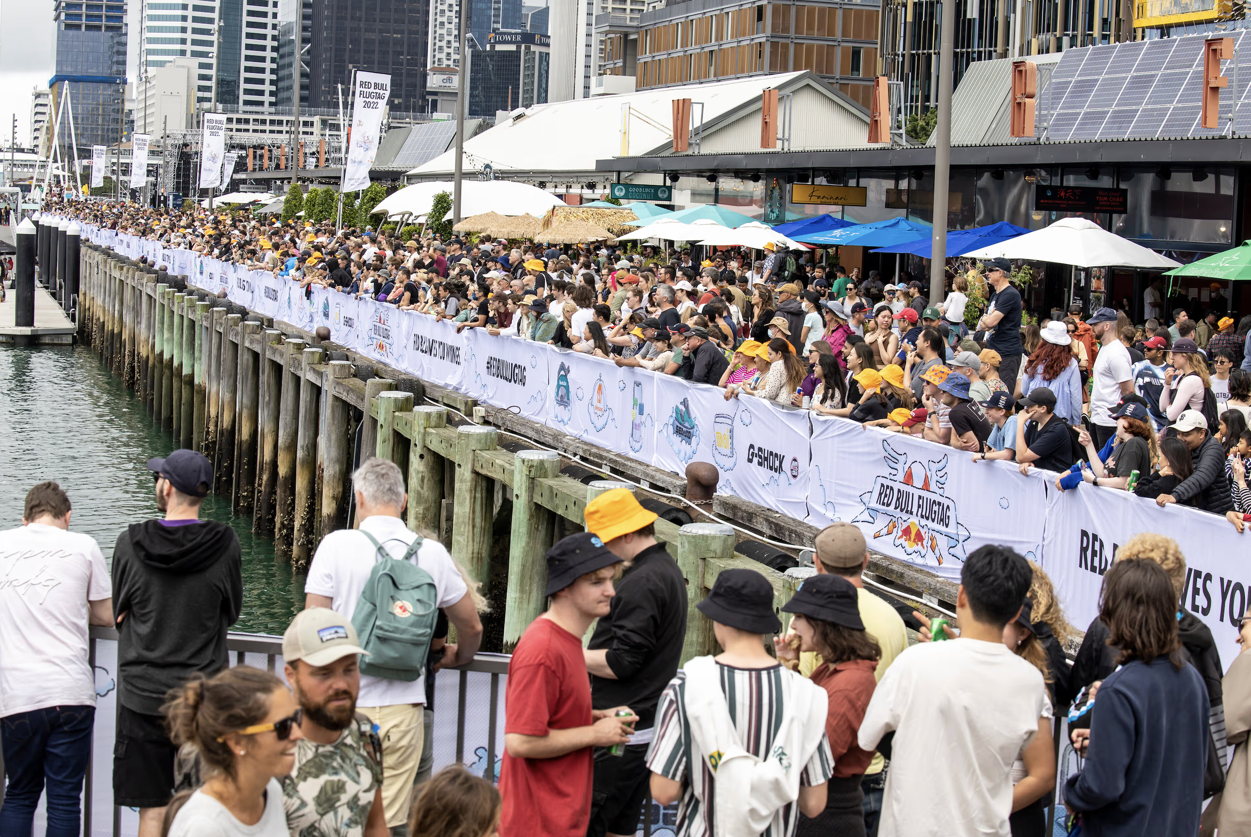A large crowd of people gathered along a waterfront, behind a white barricade with branding for Red Bull Flugtag 2022. The crowd is standing under umbrellas, with modern city buildings in the background.