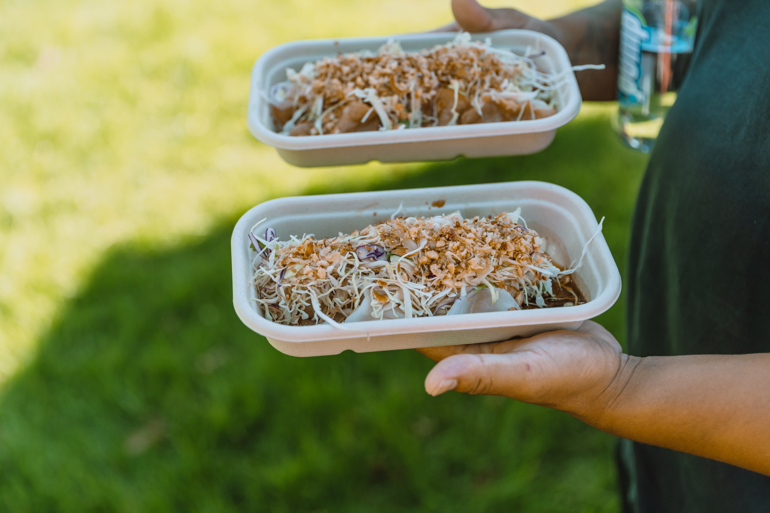 Person holding two styrofoam food containers filled with shredded meat, coleslaw, and crushed peanuts over a grassy outdoor setting.