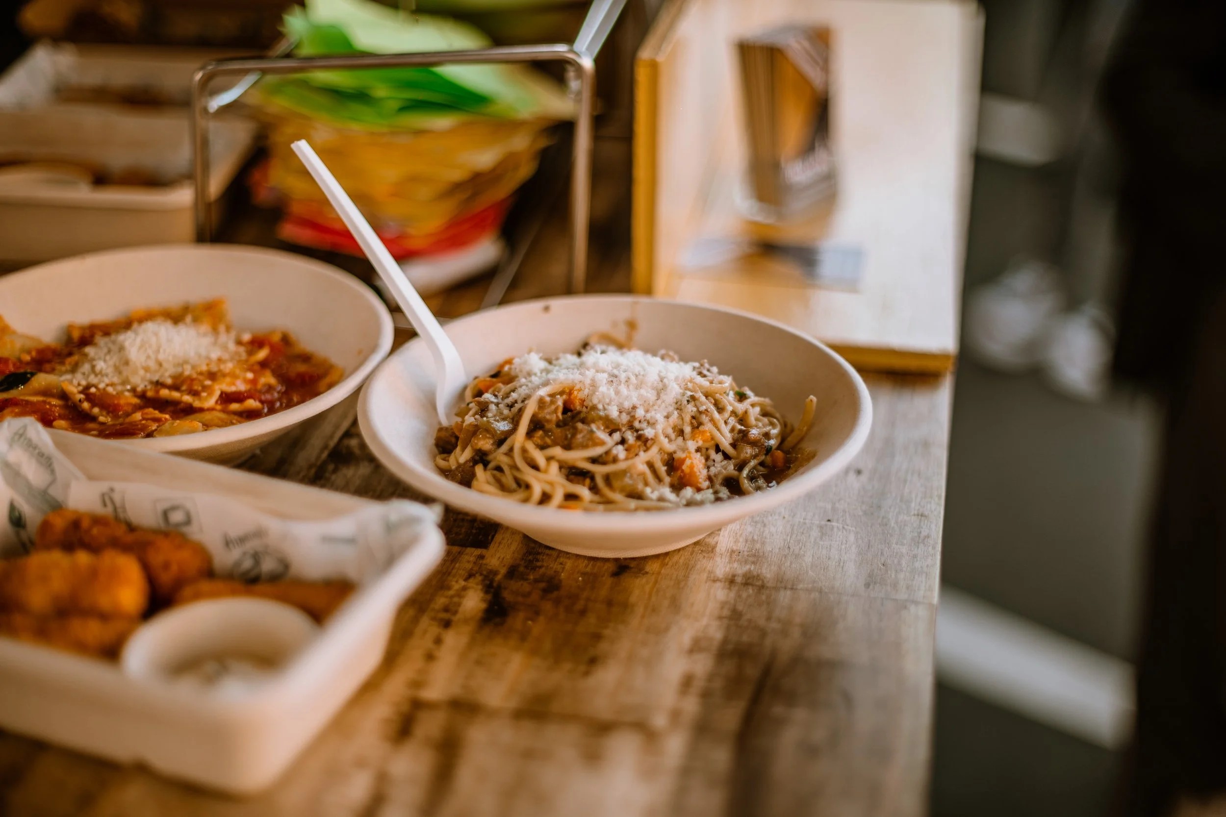 Two bowls of pasta with grated cheese, one with spaghetti and the other with a different type of noodle, on a wooden surface with a background of shelves and containers.