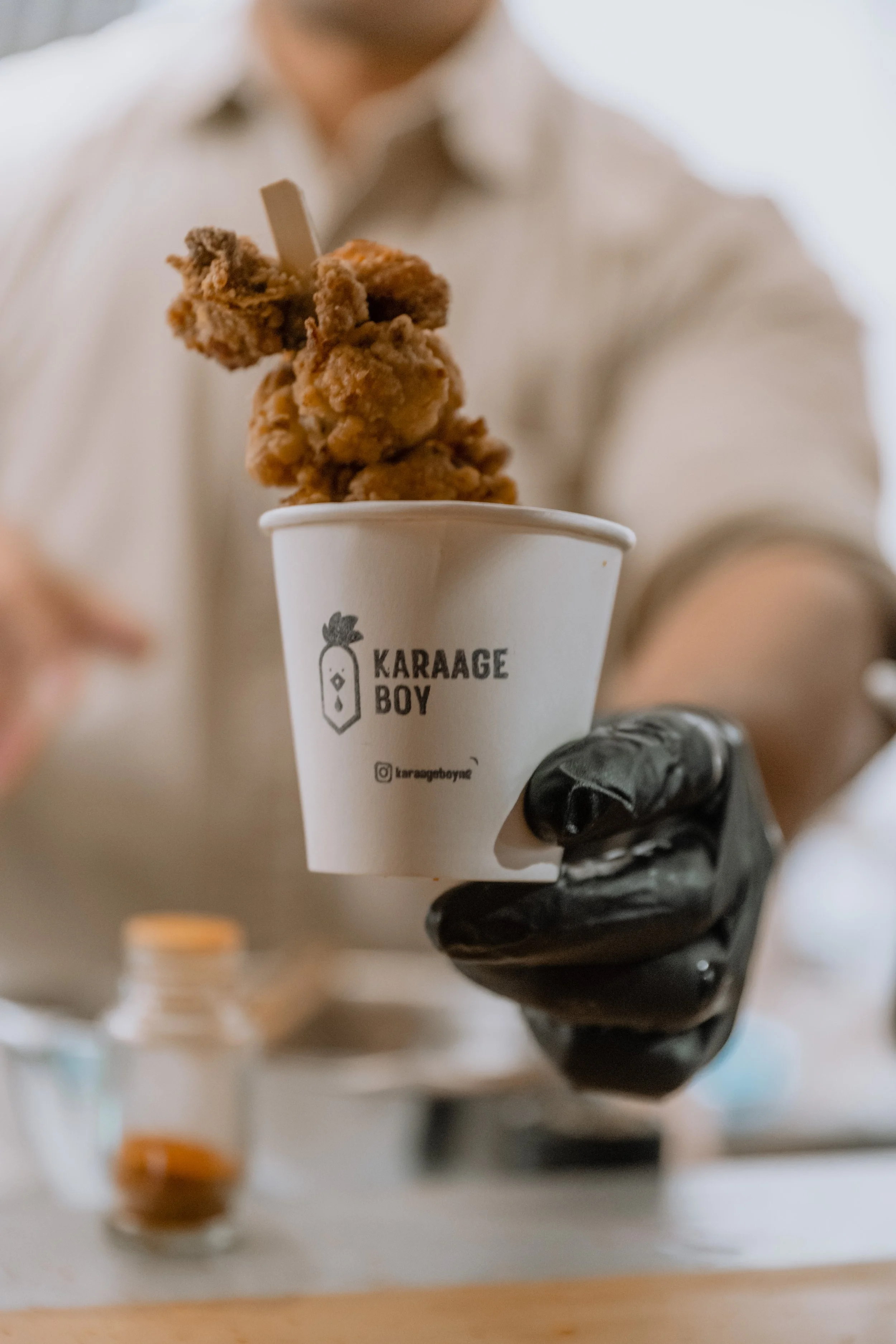 Close-up of a hand wearing a black glove holding a small white paper cup with the logo "KARAAGE BOY" and an Instagram handle. The cup contains pieces of fried chicken. In the background, blurred objects are visible.