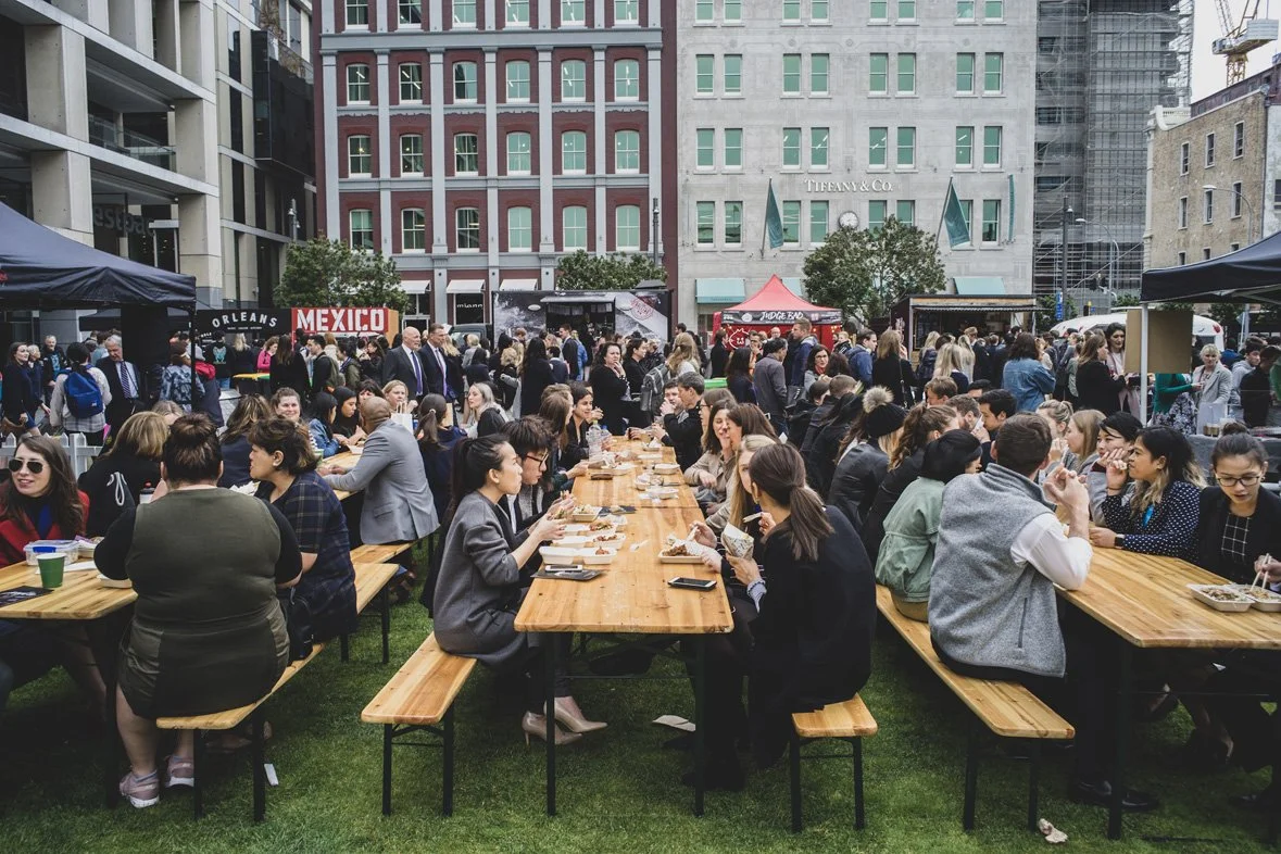 Crowd of people dining at long wooden tables outdoors in an urban setting during daytime, with buildings and tents in the background.