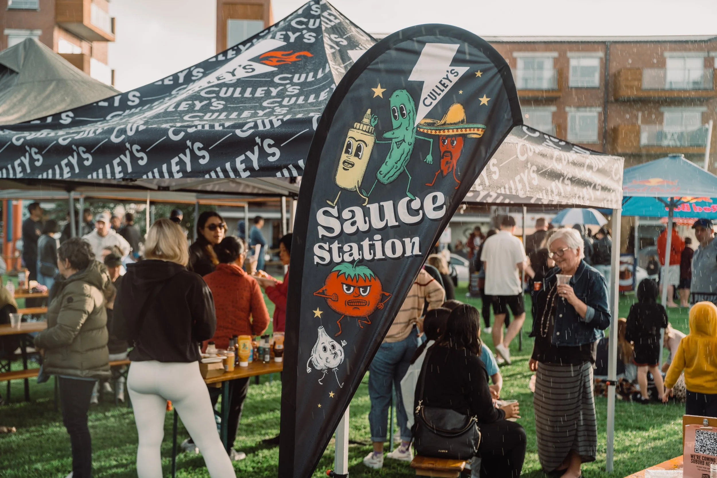 A crowd gathered outdoors under tents and flags at a festival or market. The flag prominently displays cartoon condiments including a mustard bottle, a jalapeno pepper, and a chili pepper, with the text 'Sauce Station' and 'Culley's'. People are mingling, some eating or talking, with an urban building in the background.
