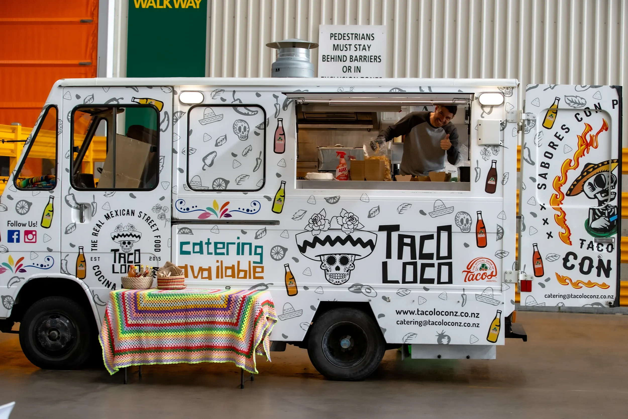 Food truck decorated with Mexican-themed graphics, including skull and chili pepper illustrations. A man inside is smiling and giving a thumbs-up. A colorful crochet tablecloth covers a small table with baskets of food in front of the truck. Signage indicates it's a taco restaurant established in 2015.