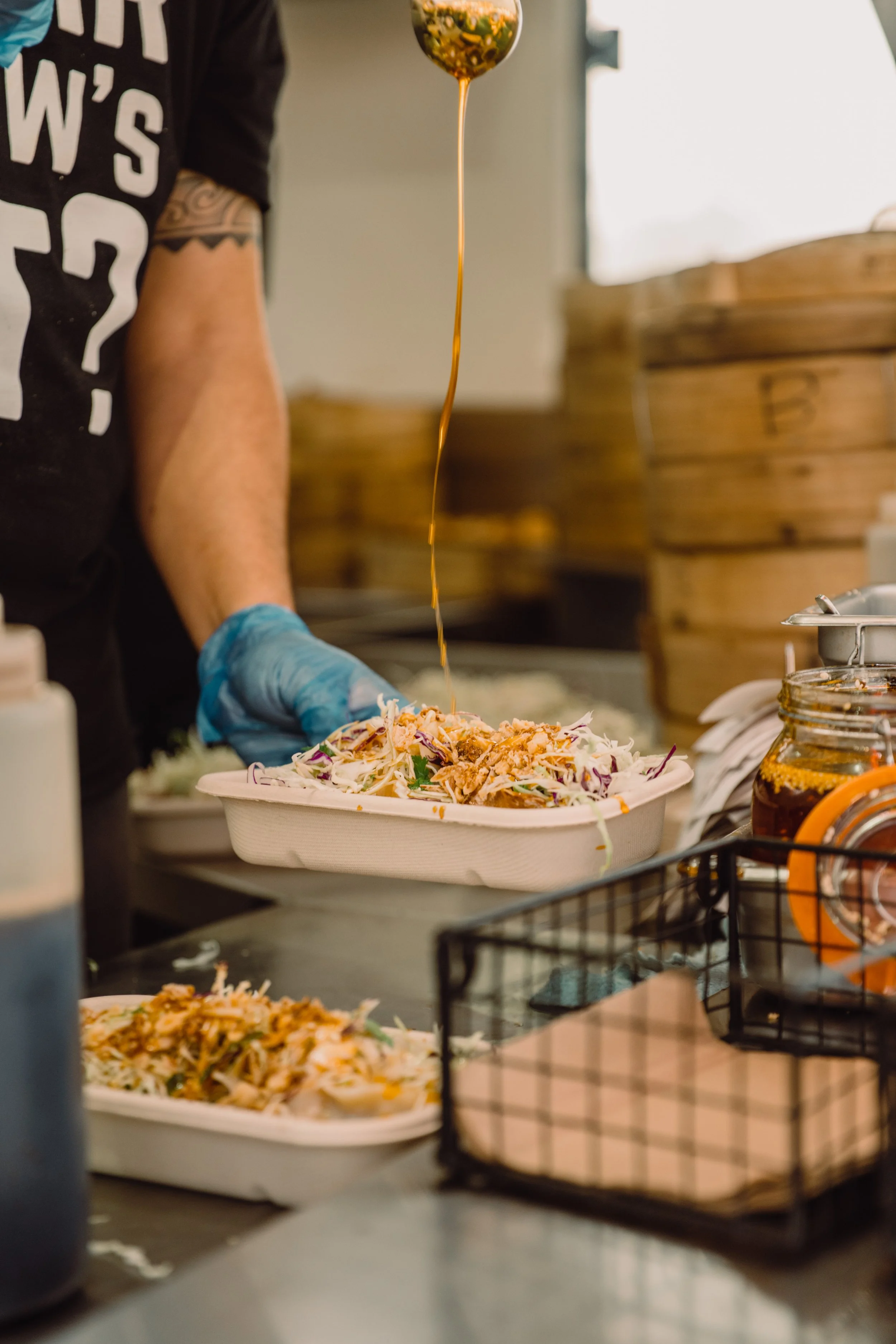 A person wearing a black shirt with white text and a tattoo on their arm is preparing food. They are pouring sauce over a salad or dish in a paper container, with jars and kitchen items visible in the background.