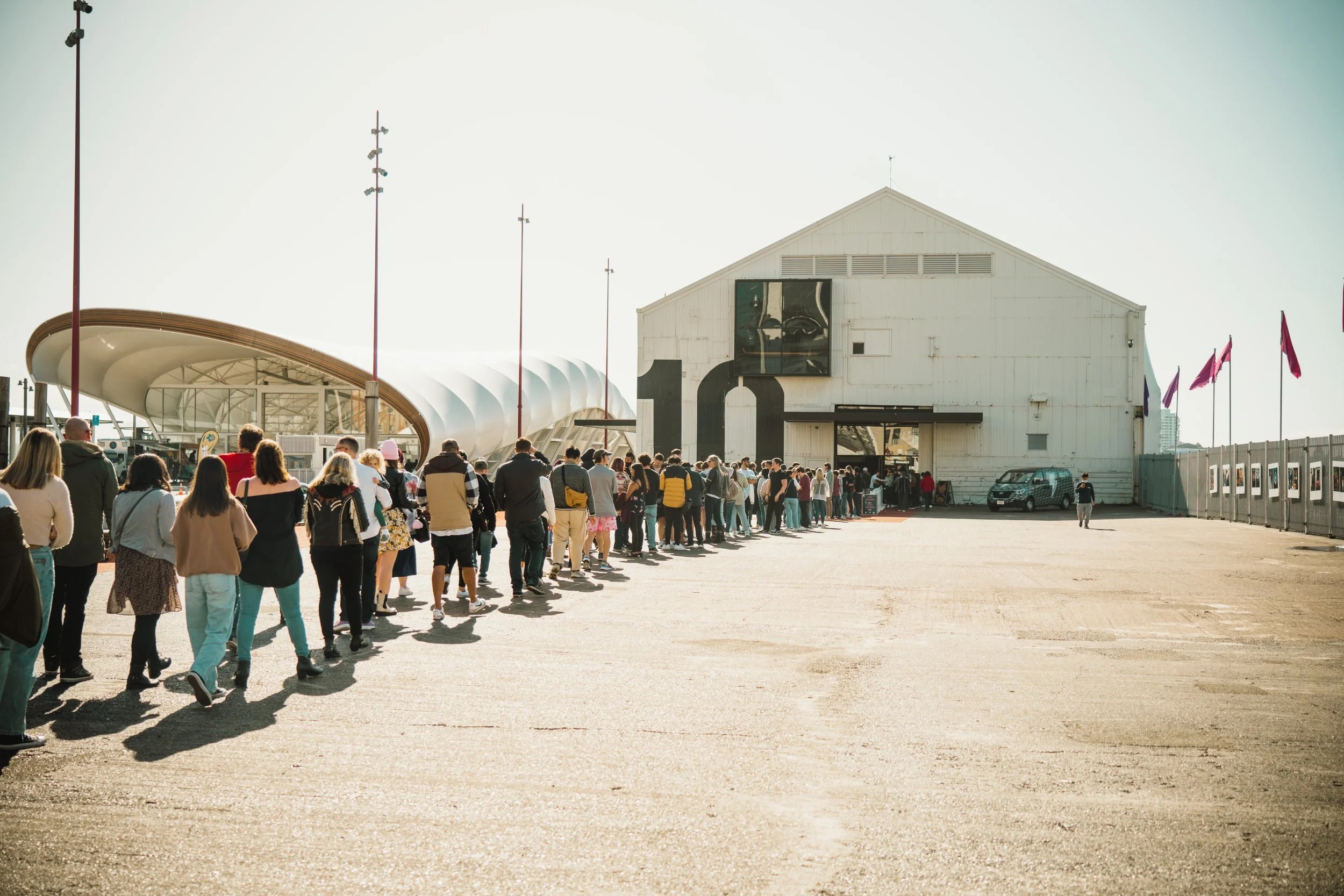 A long line of people waiting outside Shed10 with flags and modern architectural features on a clear day.