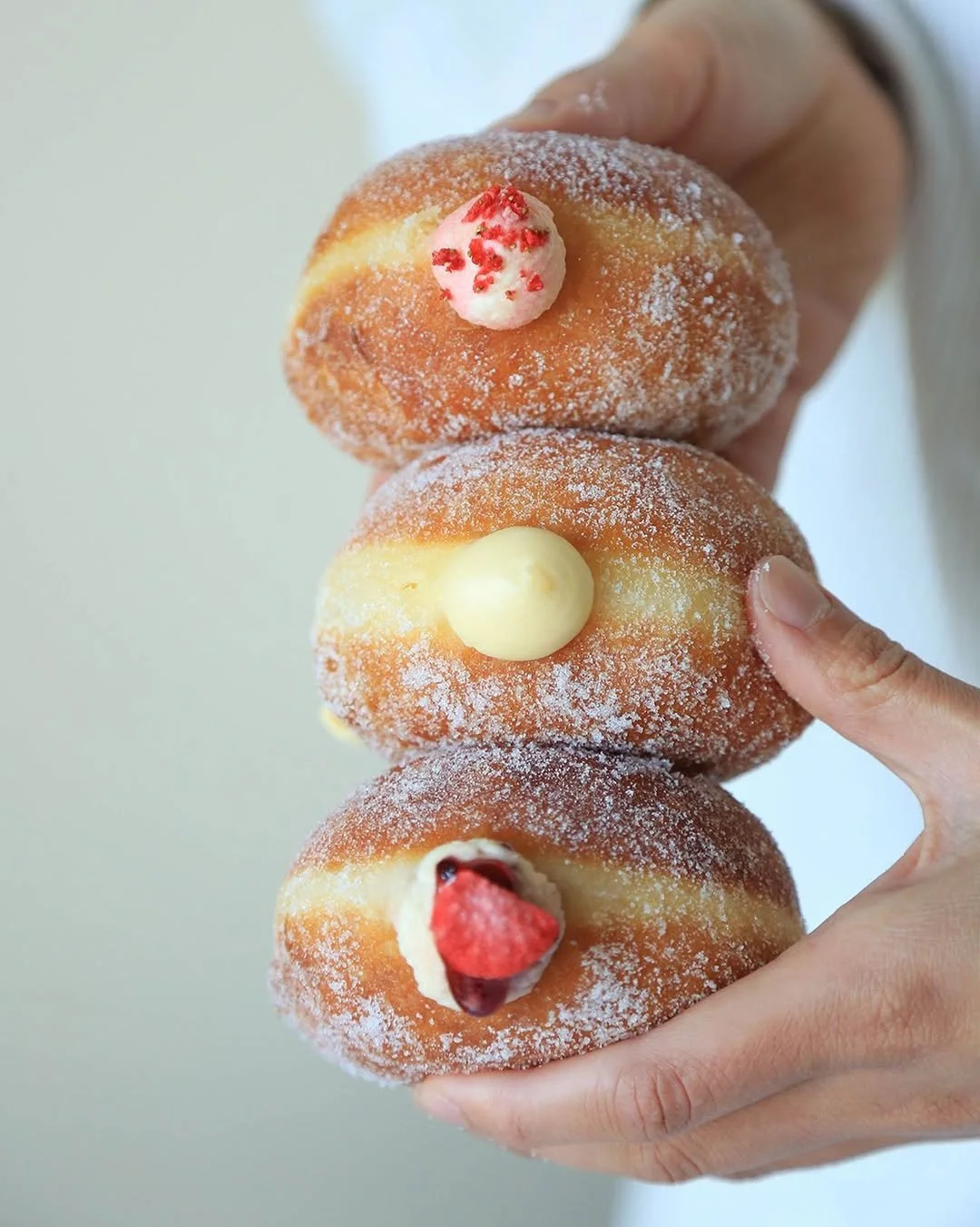 Three stacked doughnuts with powdered sugar and colorful fillings, held by a person's hand.