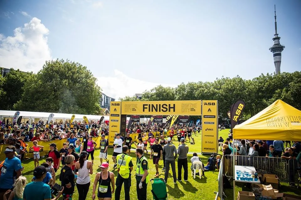 Crowd of runners and spectators at a marathon finish line, with a yellow and black banner marked "FINISH," set in a park with trees and a city skyline in the background.