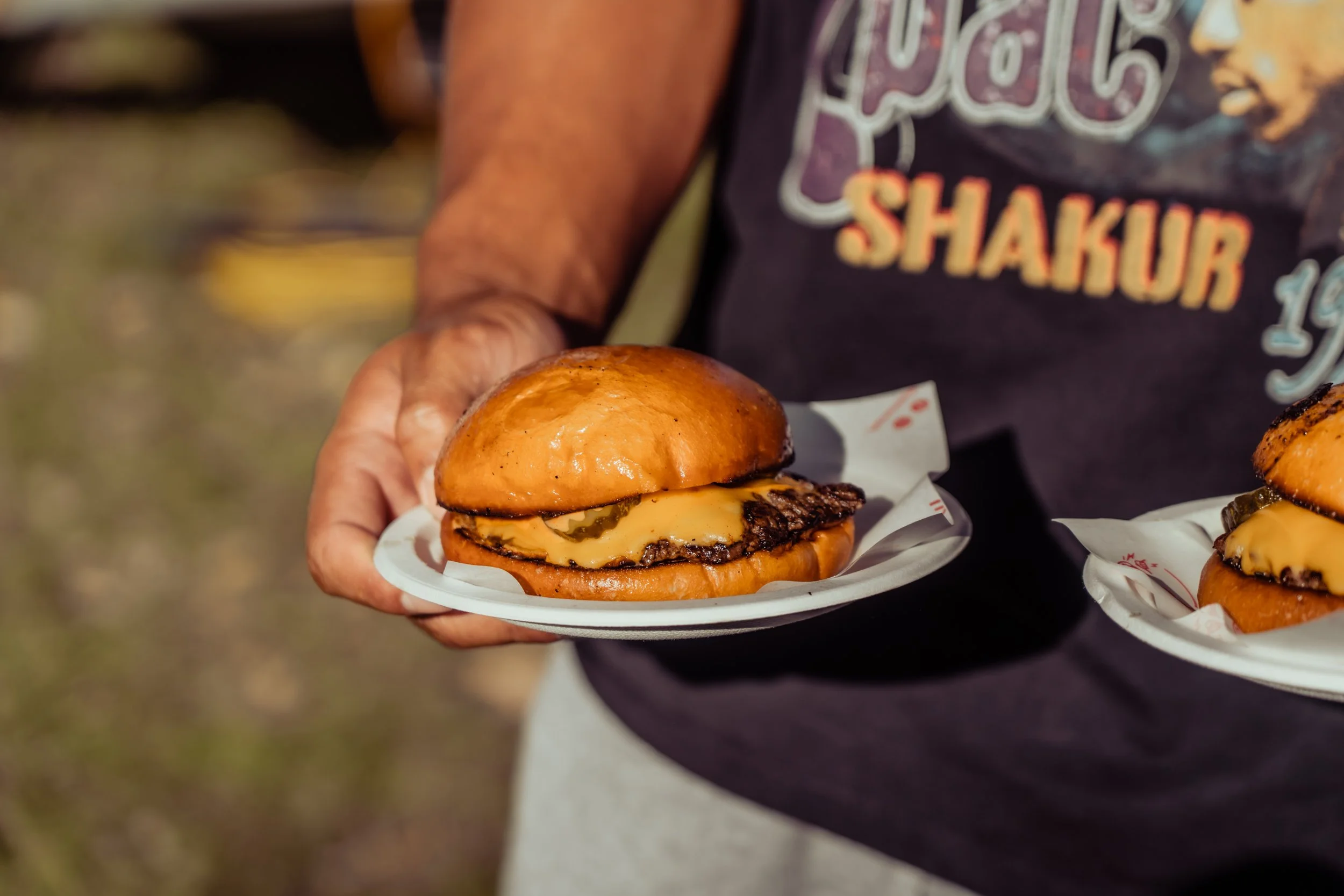 Person holding a plate with a cheeseburger, featuring a toasted bun, melted cheese, a beef patty, and pickles. The person is wearing a dark apron with some partially visible text.