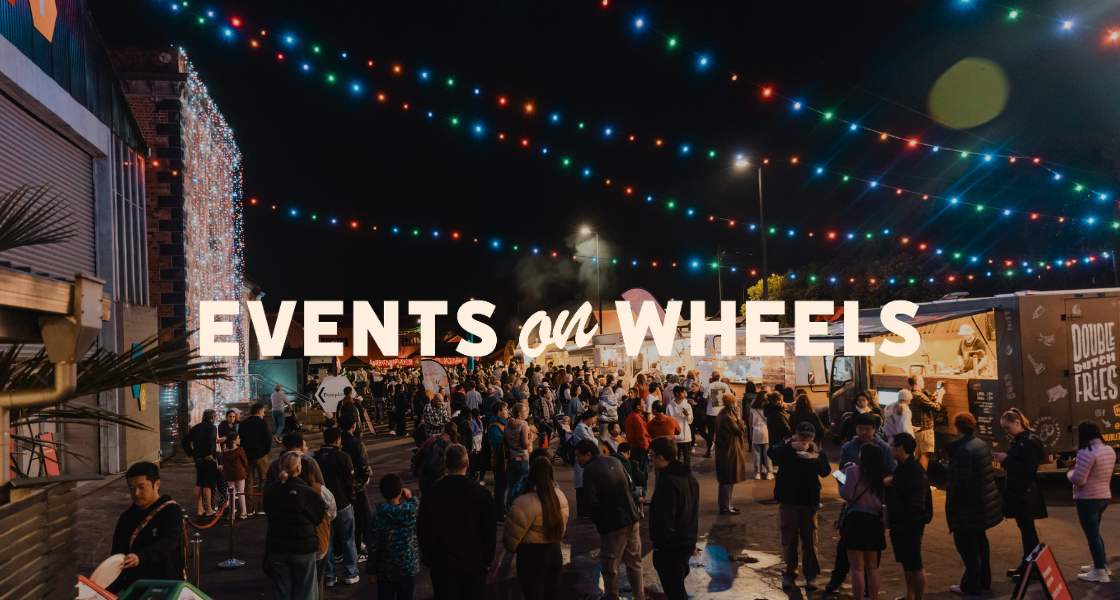 Crowd of people at night at an outdoor food market with string lights hanging above and food trucks serving food.