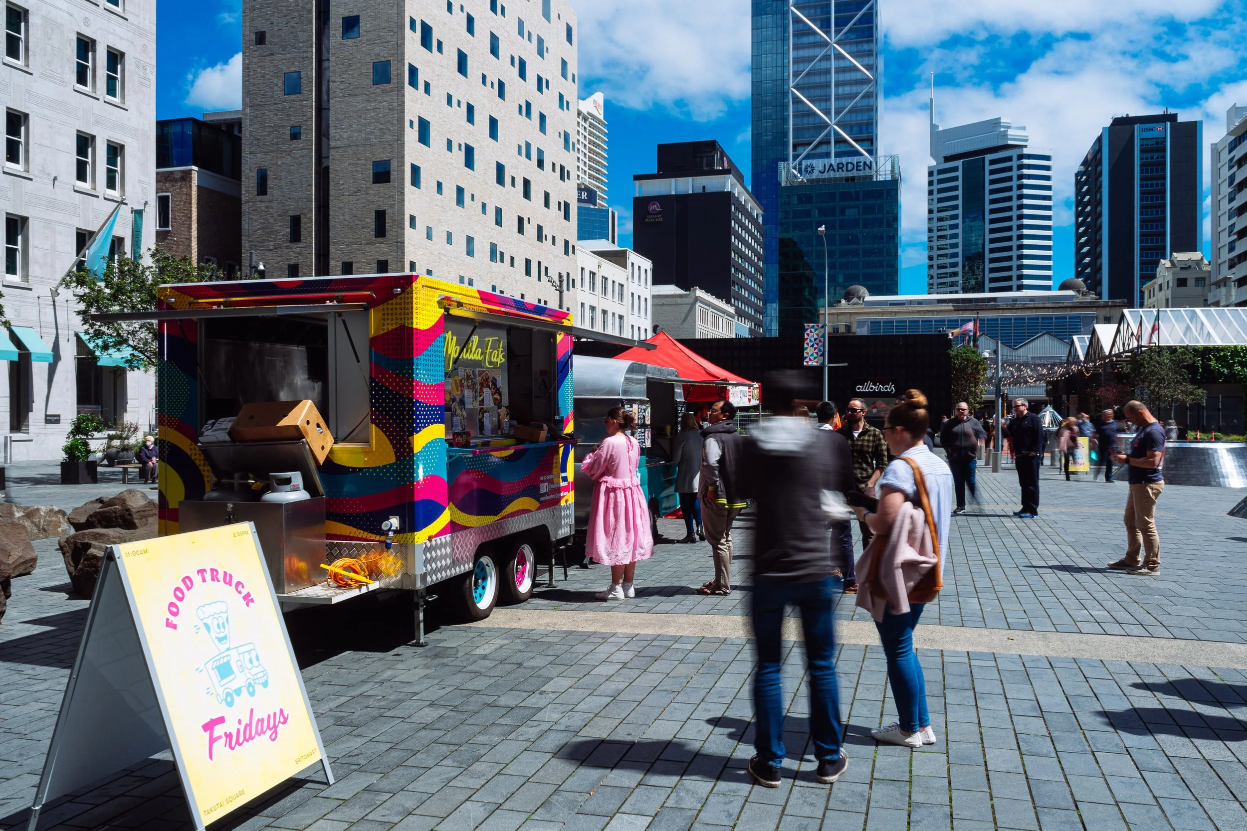 A busy city street scene with a food truck serving customers, alongside high-rise buildings and a clear blue sky. People are walking and socializing in the area.