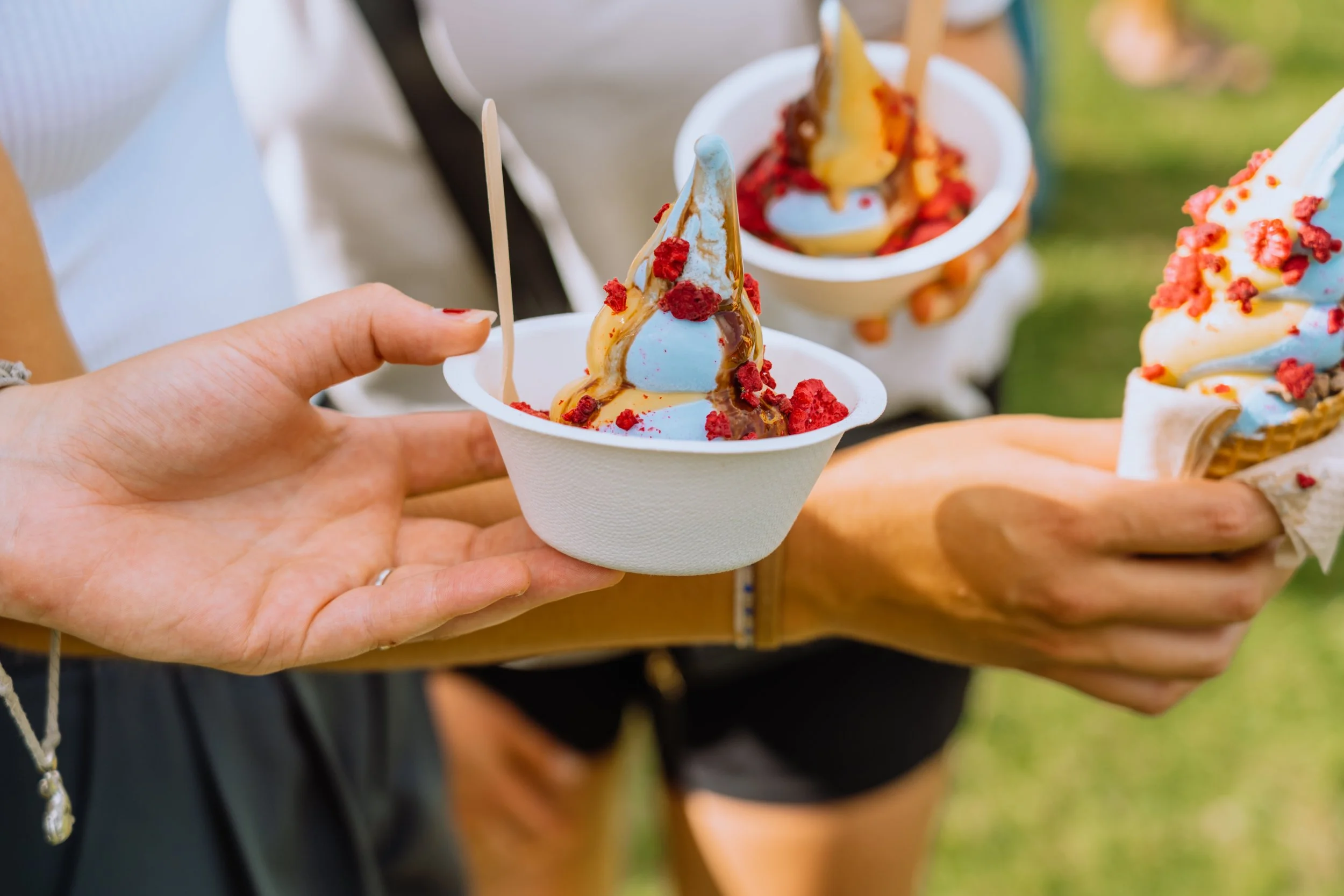 Person holding a cup of ice cream with toppings including red crumbs and caramel drizzle, with other similar cups in the background.