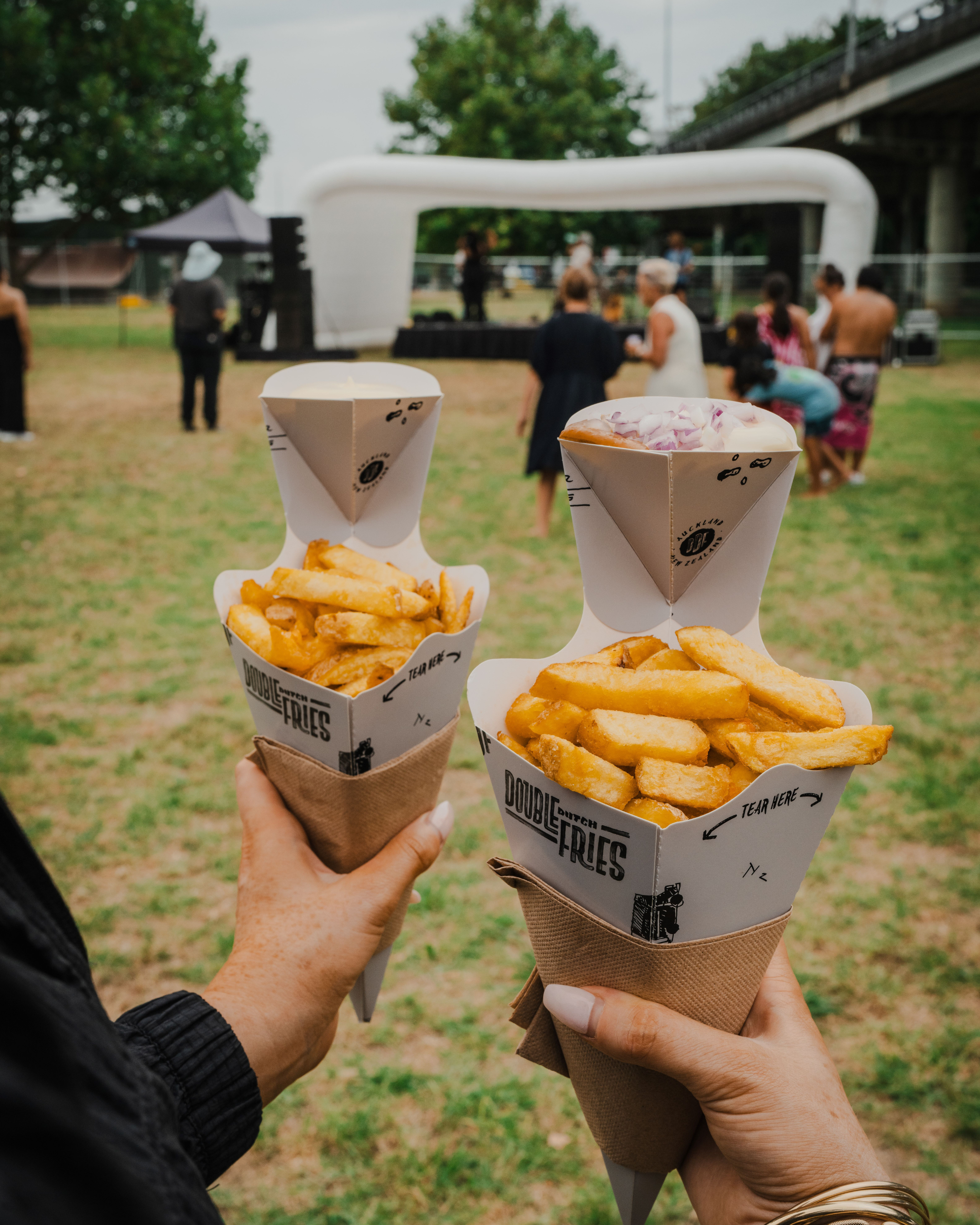 Two people holding food containers with fries and dipping sauces at an outdoor event, with a stage and attendees in the background under a cloudy sky.