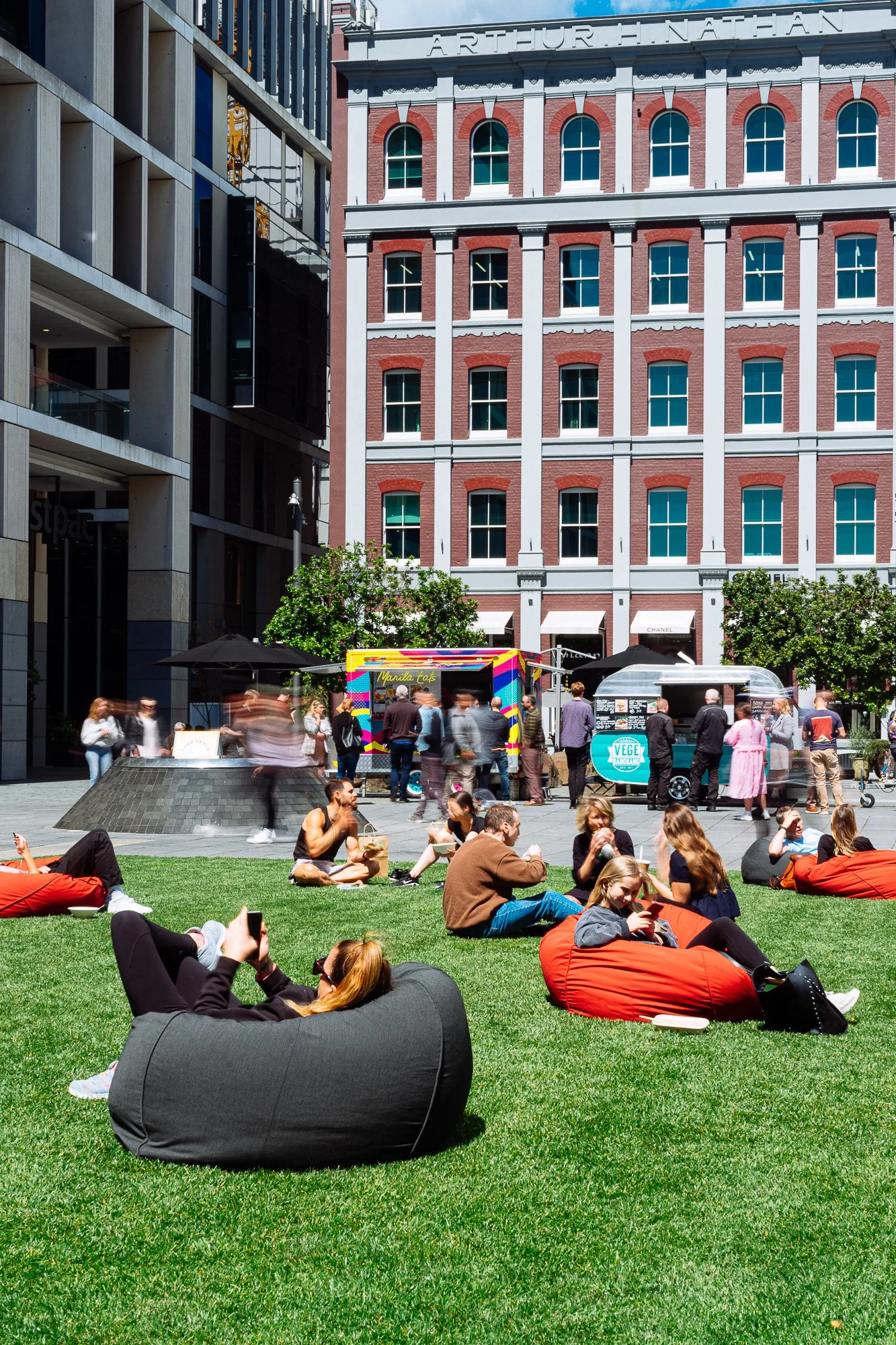 People relaxing and socializing on a grassy outdoor space in front of modern and historical buildings, with food trucks and trees.