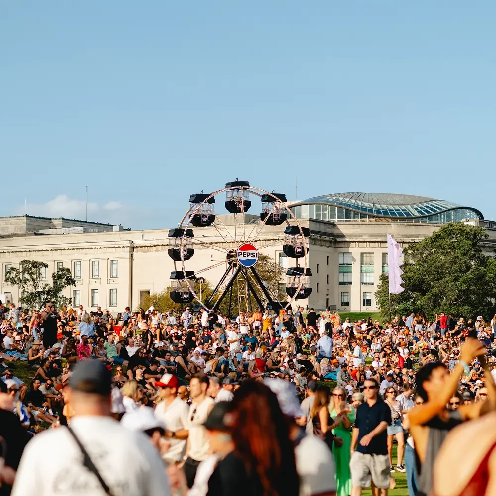 Crowd of people gathered outdoors at a festival with a Ferris wheel branded Pepsi and a building with a glass roof in the background.