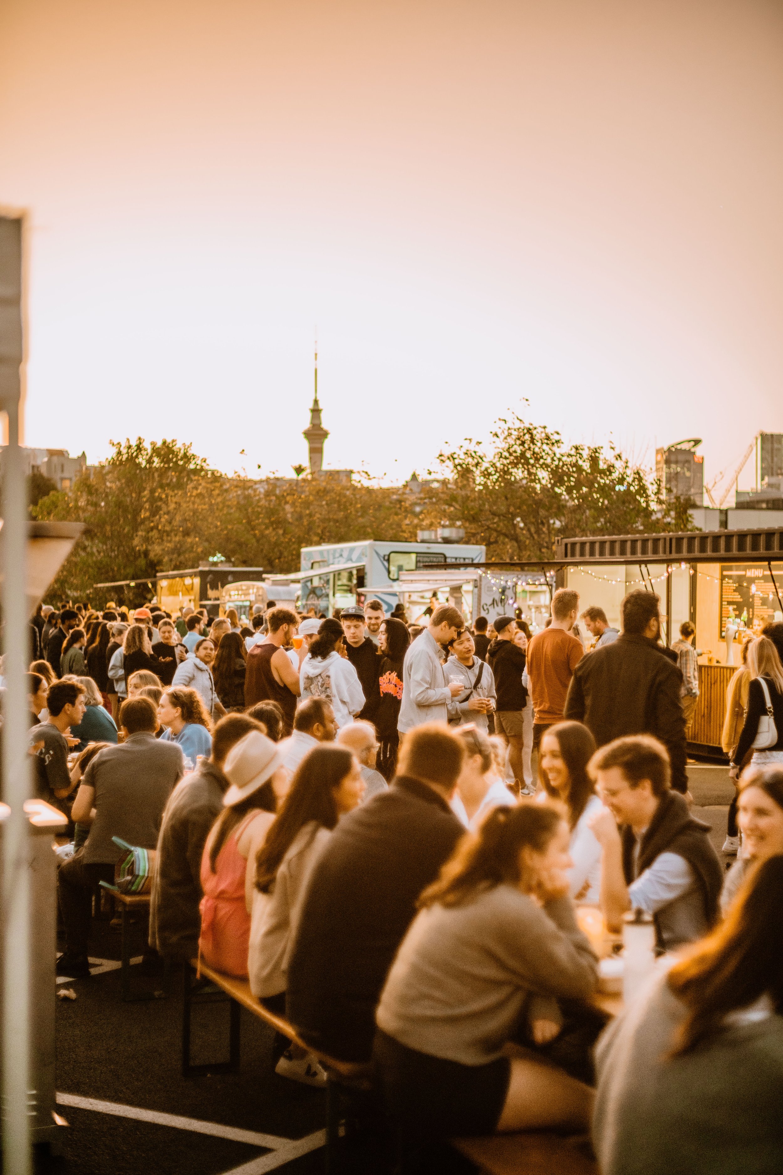 A crowd of people gathered at an outdoor event during sunset, with food trucks and trees in the background.