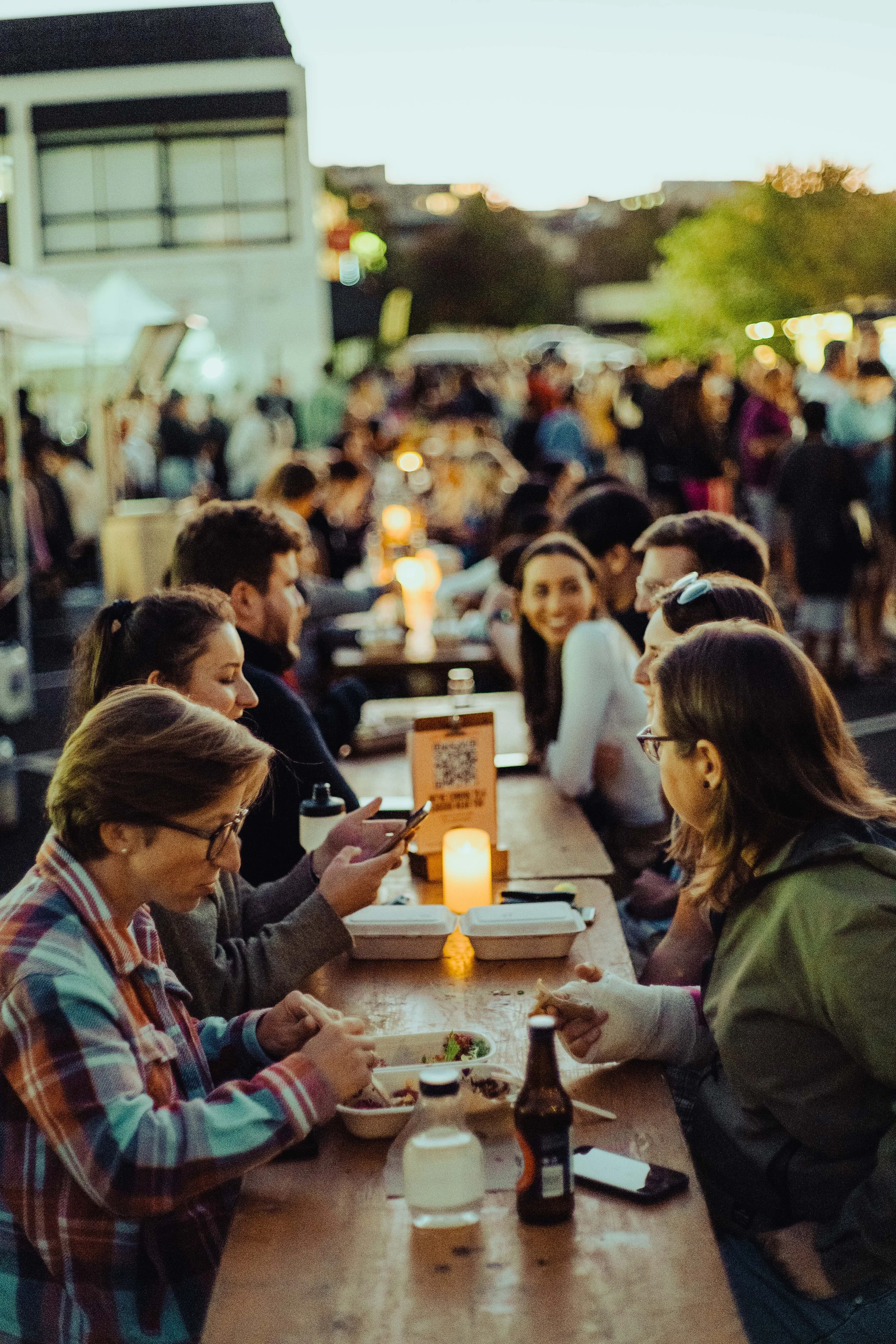People sitting at a long outdoor table dining and socializing during the evening. Some are using their phones, and there are food containers, a bottle, and a candle on the table. The background shows a blurred crowd and buildings.