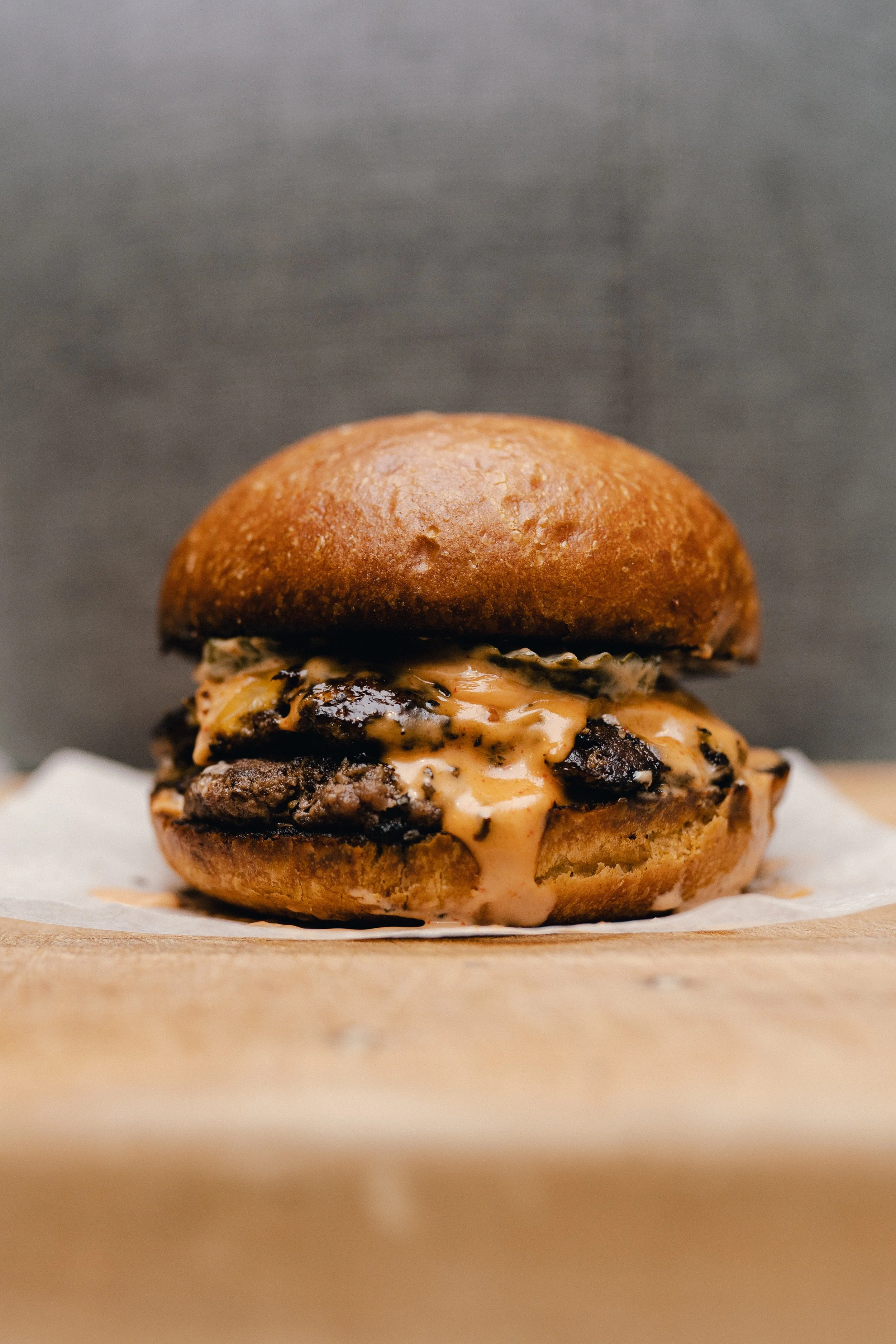 A close-up of a gourmet burger with a toasted bun, melted cheese, a beef patty, and sauce, placed on a white paper on a wooden surface.