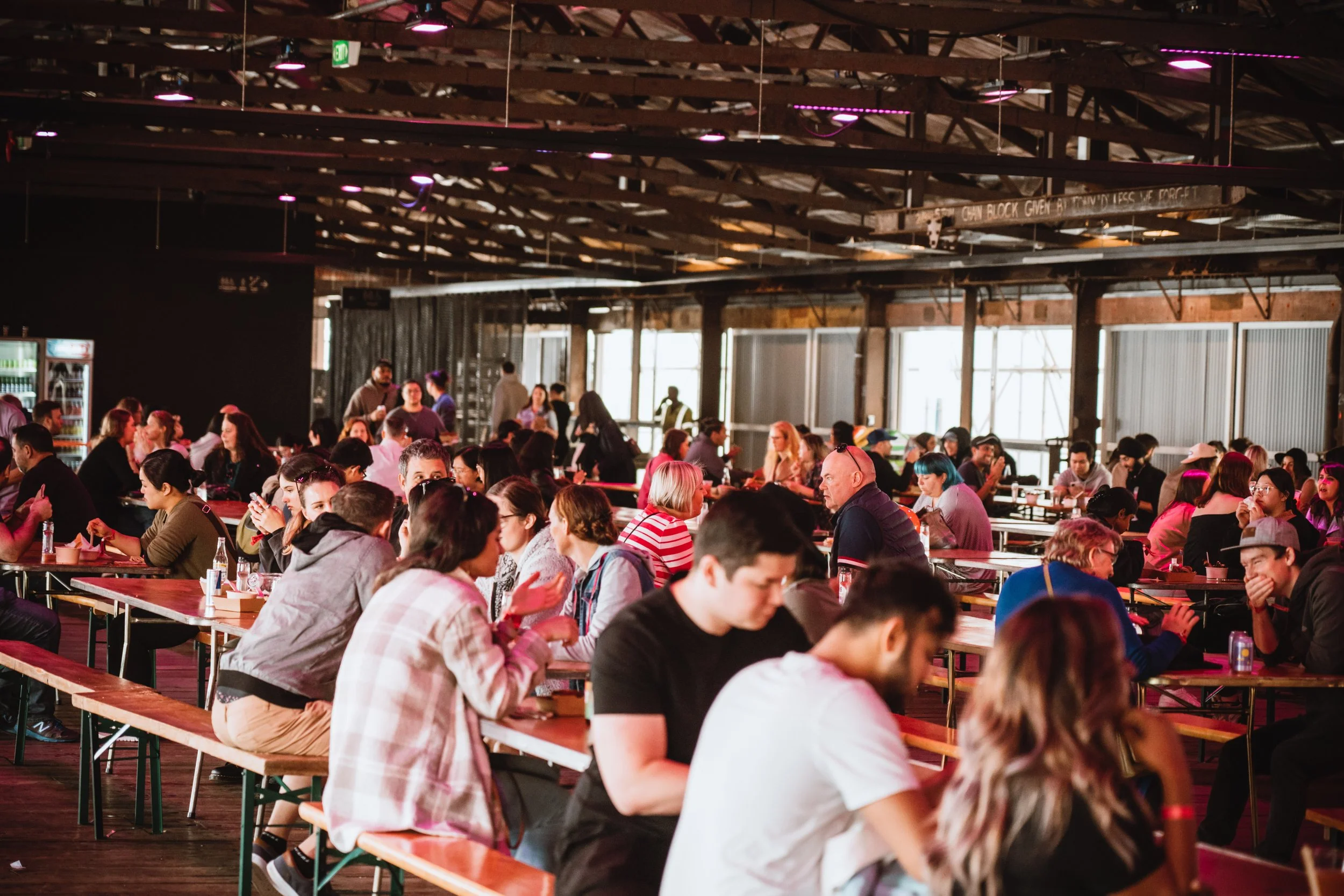 People sitting at tables in a large, industrial-style indoor space with wooden ceiling beams and large windows.