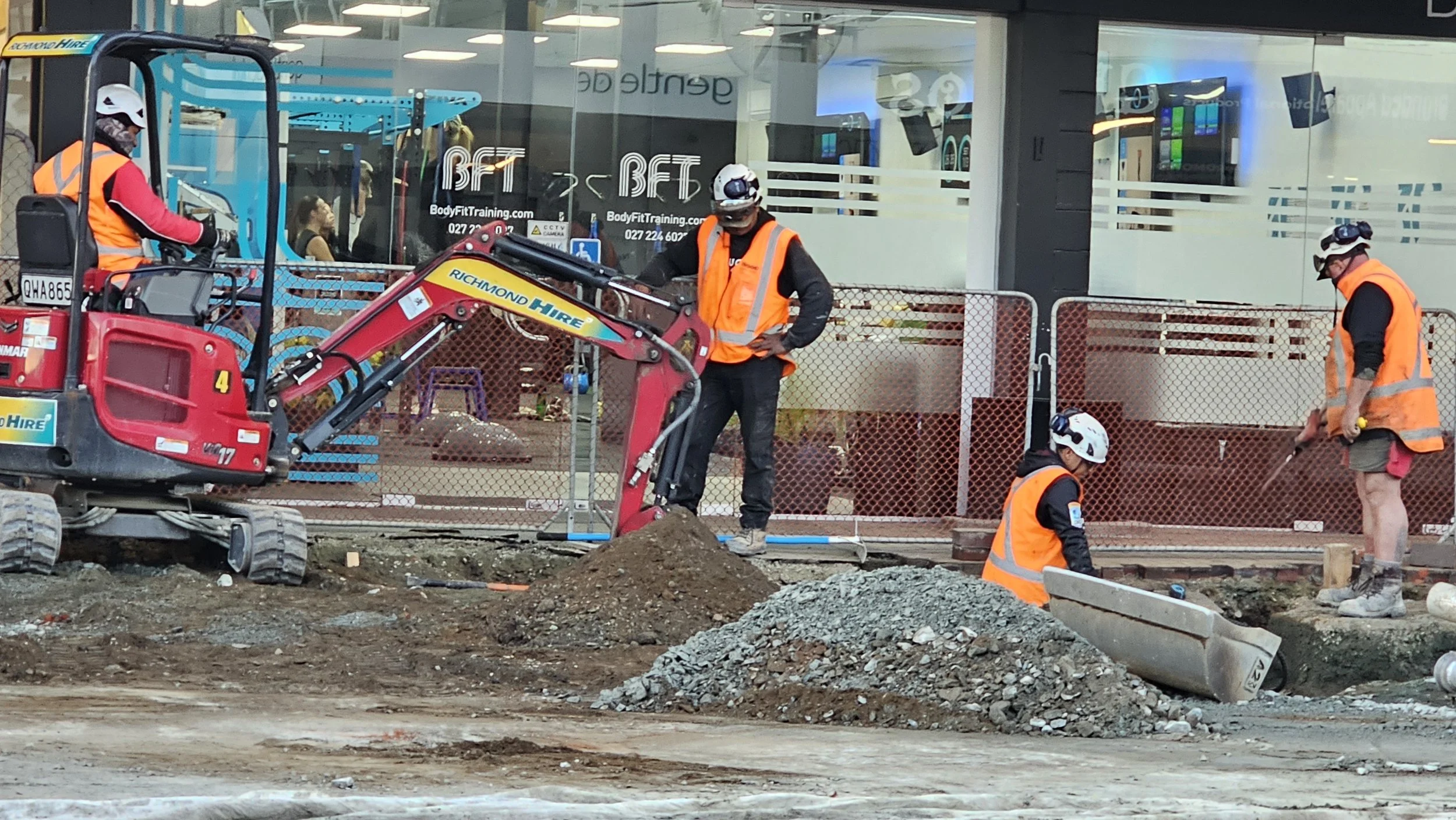 Construction workers in orange safety vests and helmets working on a sidewalk or road reconstruction project in front of a building with large glass windows.