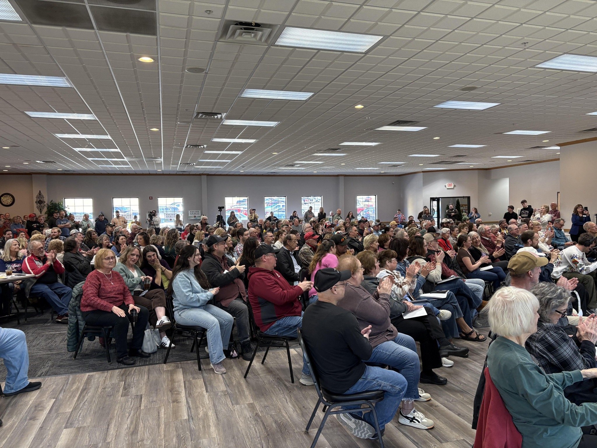 A large crowd of people seated and standing in a spacious indoor hall, attending an event or presentation, with some clapping and engaging with the speaker.
