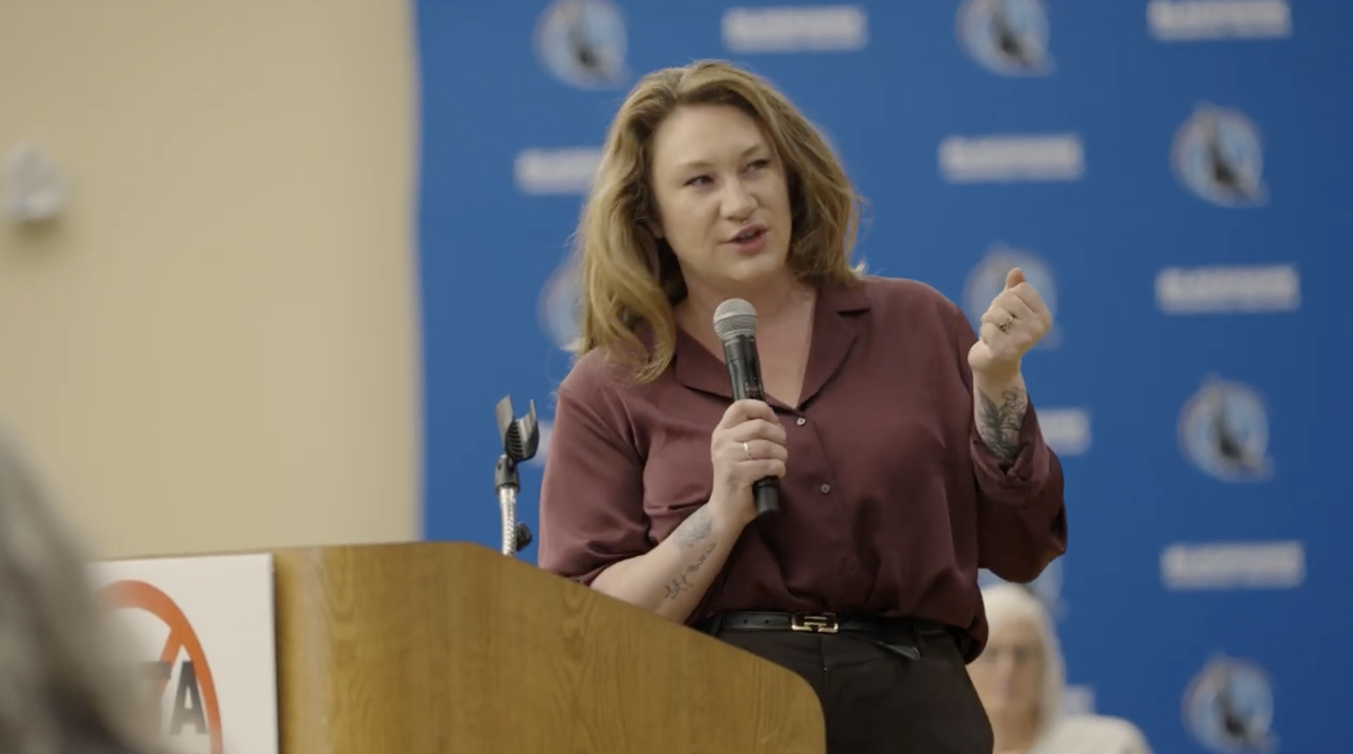A woman with shoulder-length red hair, wearing a maroon shirt, is speaking into a microphone at a podium during a press conference or event. She appears to be making a point with her clenched hand. There is a blue backdrop with a logo behind her.