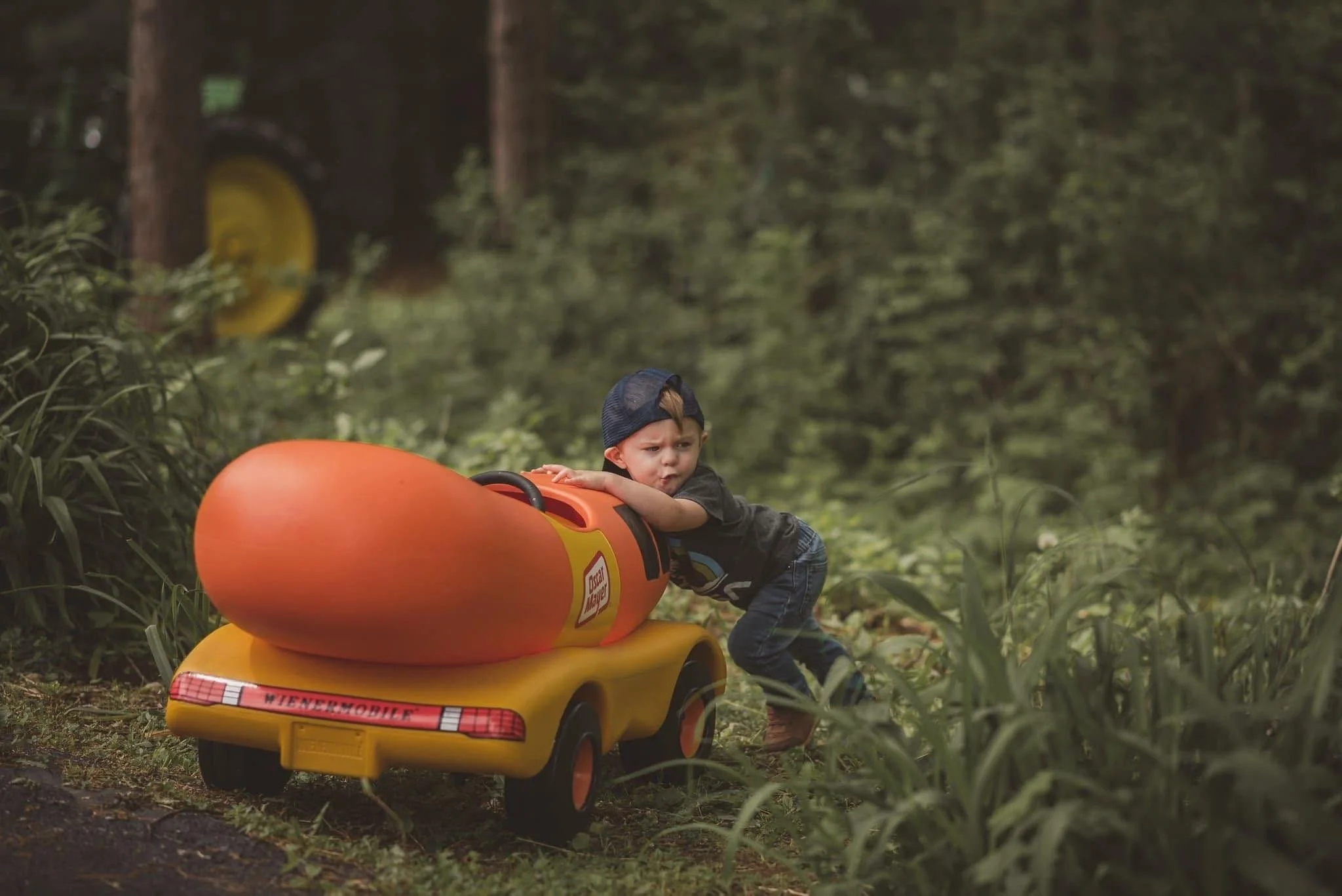 A young boy in a baseball cap pushes a toy yellow and orange car with a rocket on the back through a forested area.