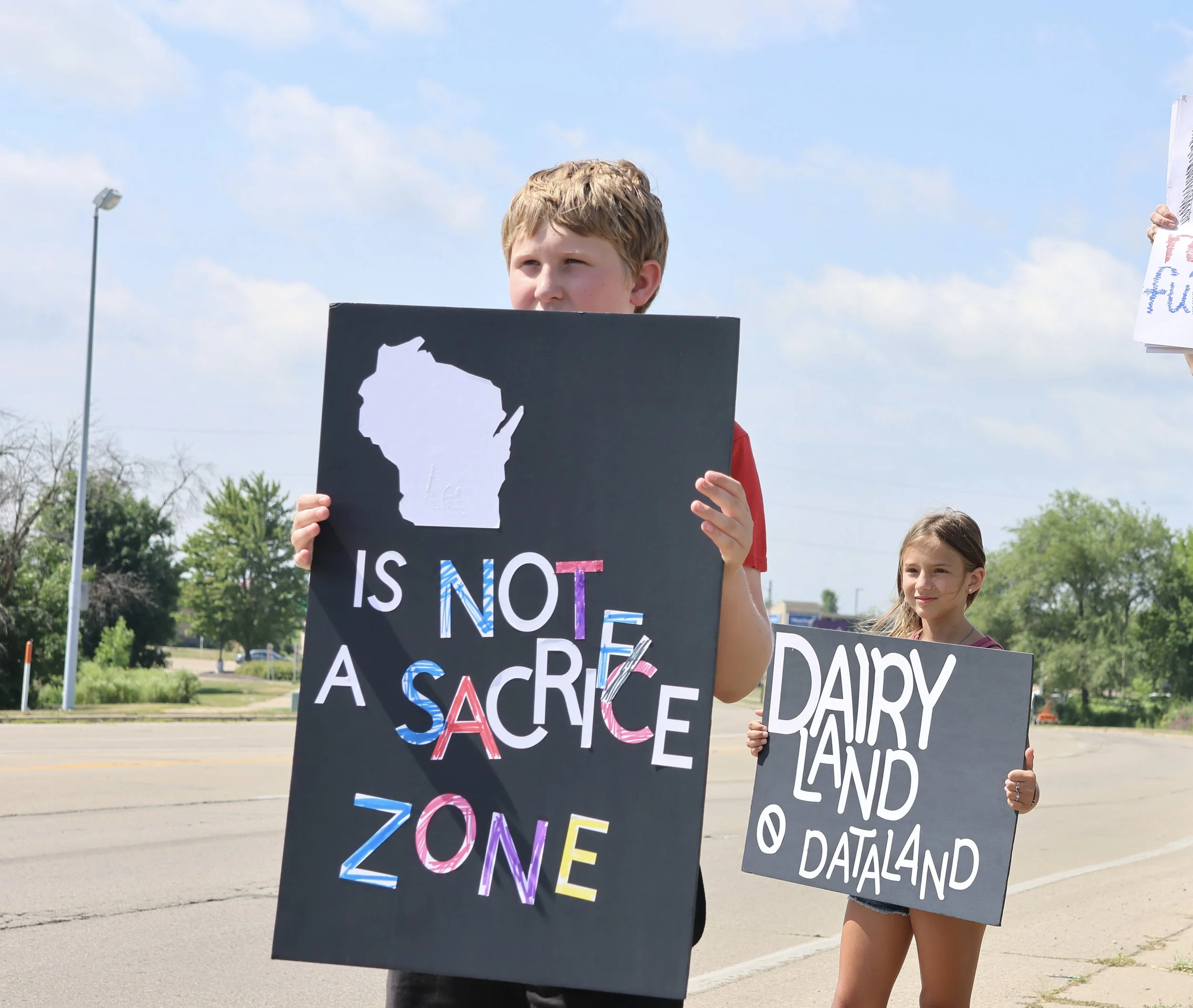 Teenagers holding protest signs outdoors on a sunny day with a blue sky. One sign reads 'ZONE' with colorful letters, another partially visible sign mentions 'DREAM' and 'SACRIFICE.'