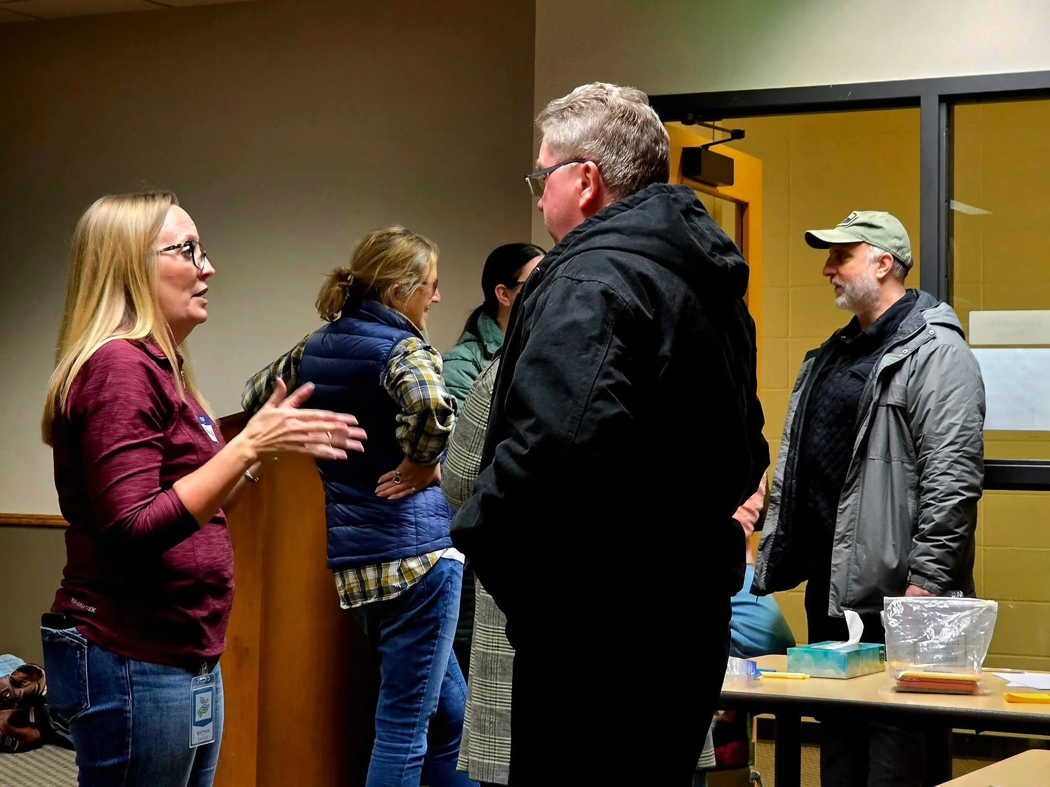 People talking at an indoor event, standing in a queue or line. A table with supplies is visible on the right.