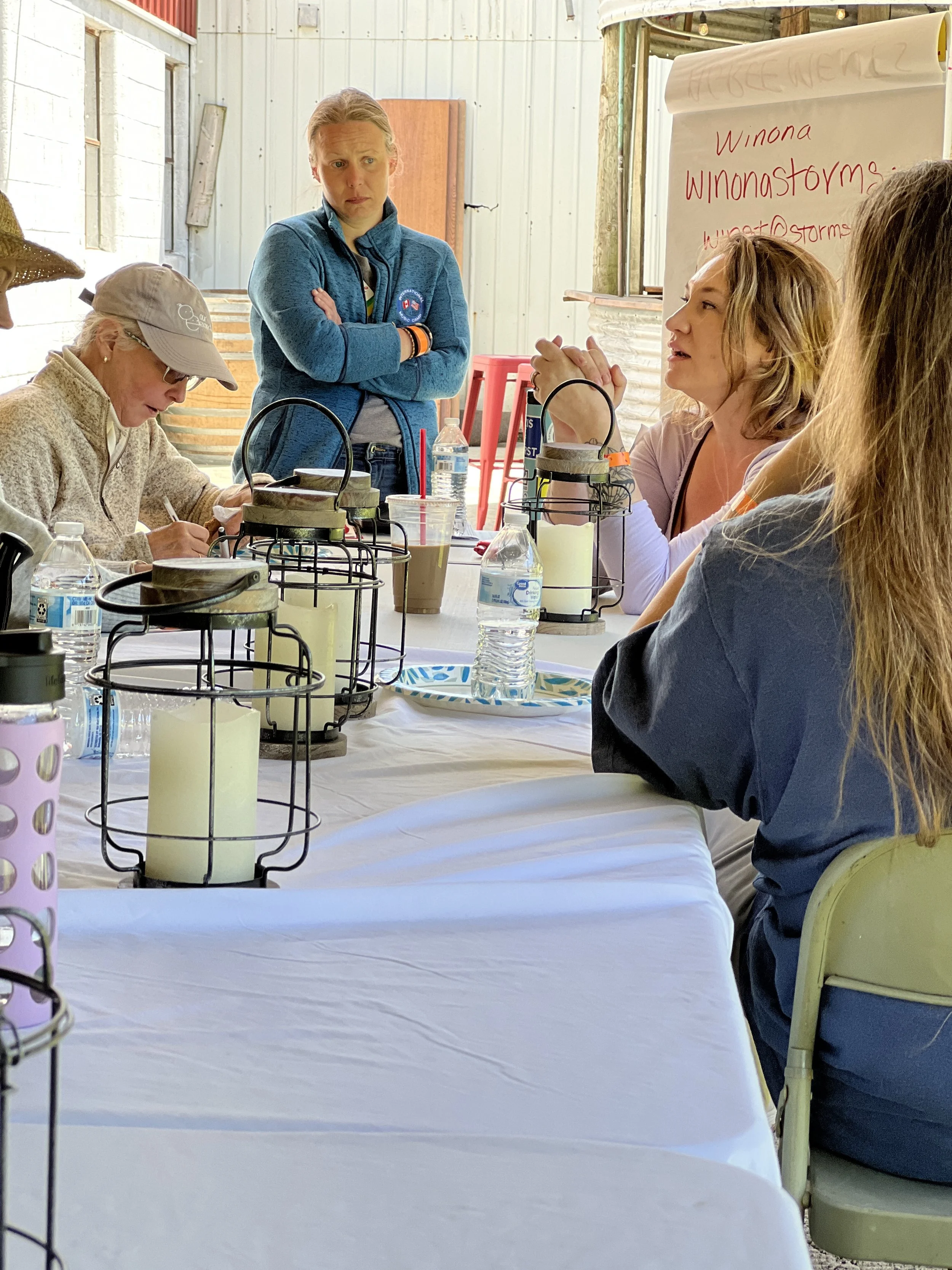 Four women sitting at a long table with a white tablecloth, engaging in a discussion. One woman is standing with her arms crossed, looking at the speaker. The table has water bottles, coffee cups, and candles in lanterns. There is a flip chart with handwritten notes in the background.