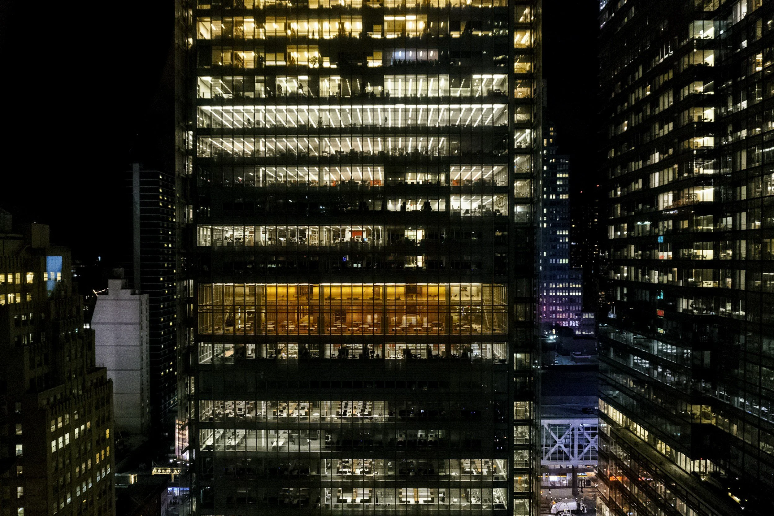 Nighttime view of a tall office building with illuminated interior through glass walls, showing multiple floors with visible offices and workspaces.