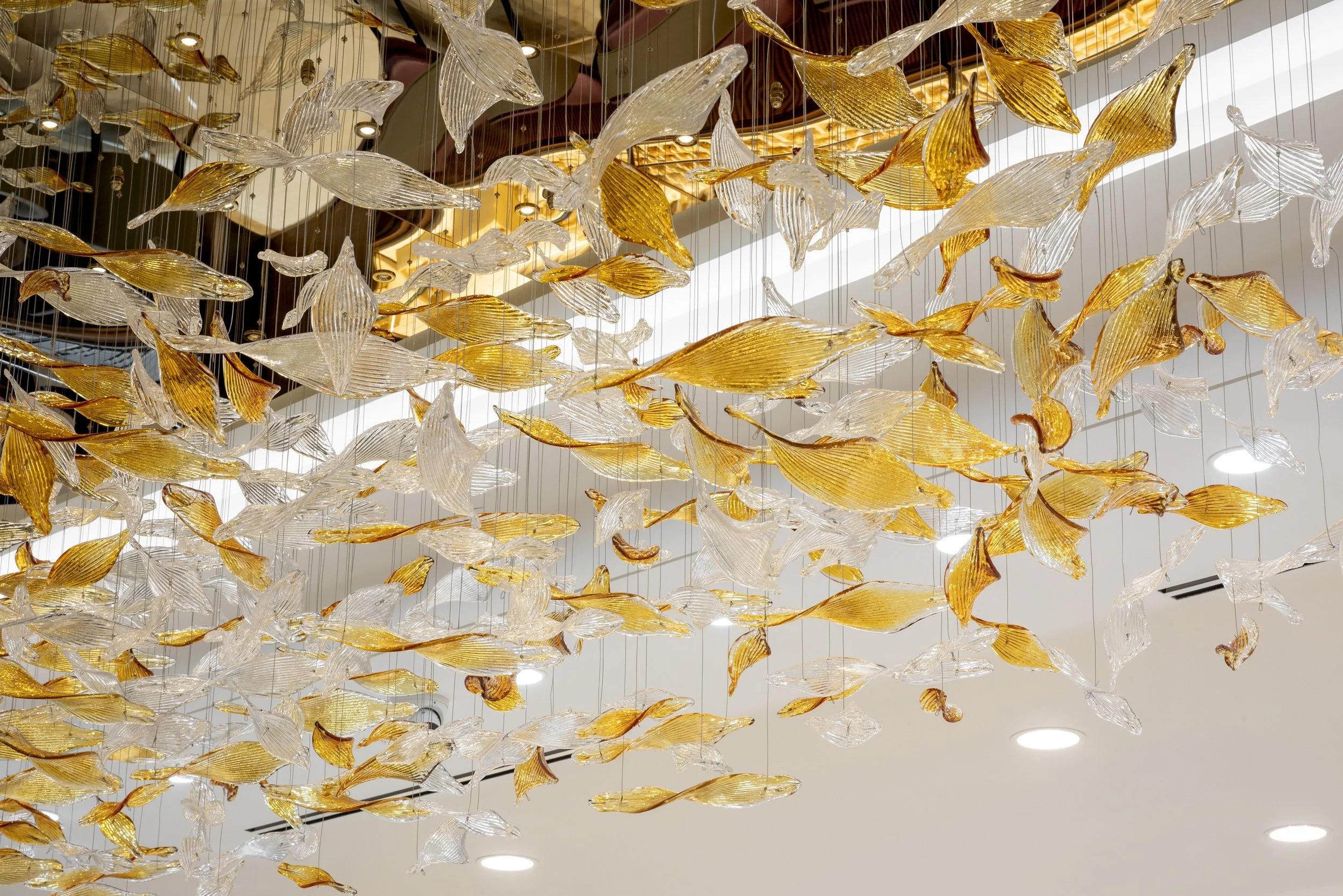 A large, artistic chandelier made of multiple glass pieces resembling fish or leaves, with gold and clear glass accents, hanging from the ceiling of an indoor space.