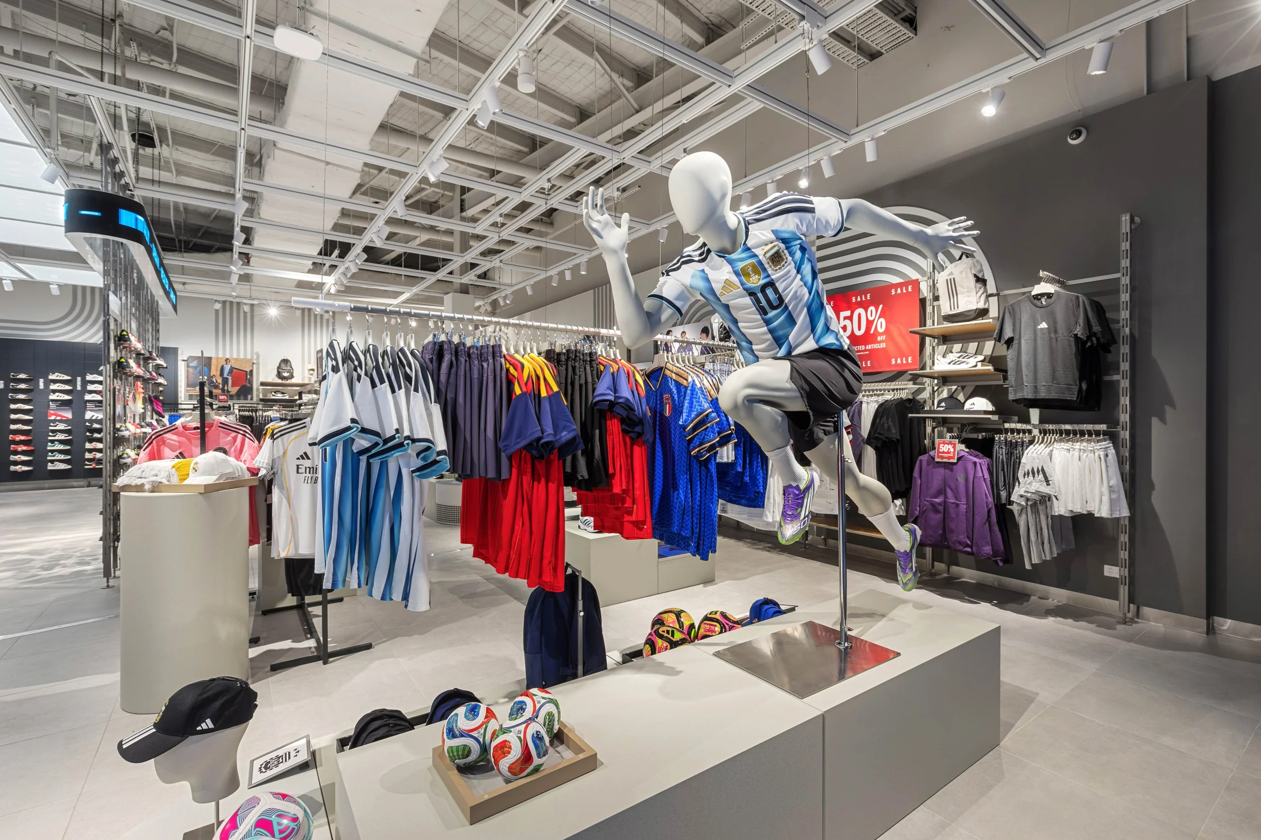 A mannequin wearing a soccer jersey, shorts, and soccer shoes in a sports apparel store display. The display includes soccer balls, hats, and clothing racks with jerseys and other sportswear, with a sale sign indicating 50% off.