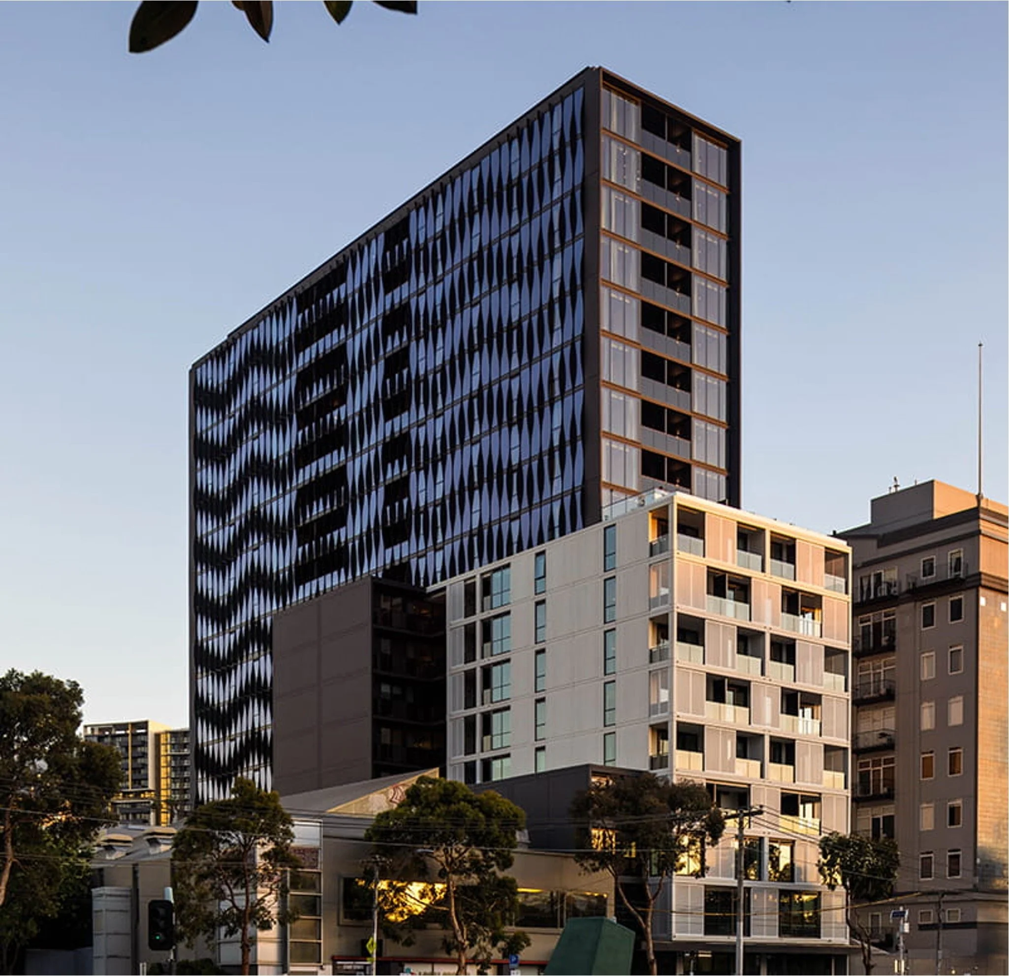 A modern high-rise building with a distinctive black exterior featuring diamond-shaped patterns, situated in an urban area with other buildings and trees visible at the ground level, illuminated by natural daylight.