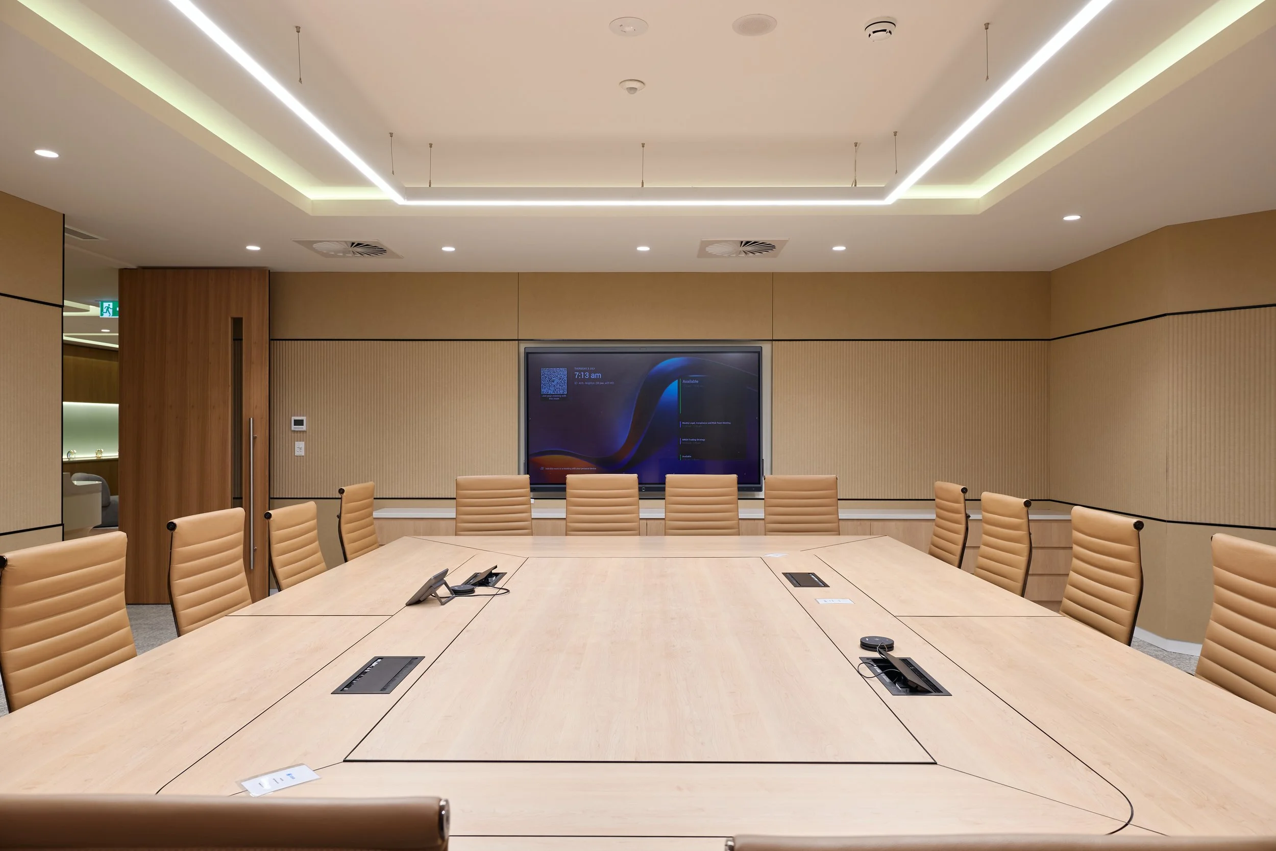 A modern conference room with a large oval wooden table surrounded by tan leather chairs. The room features beige walls, a large digital screen on the back wall, and a ceiling with integrated lighting and ventilation systems. There are telephones on the table and a few pieces of paper clipped to the chairs.