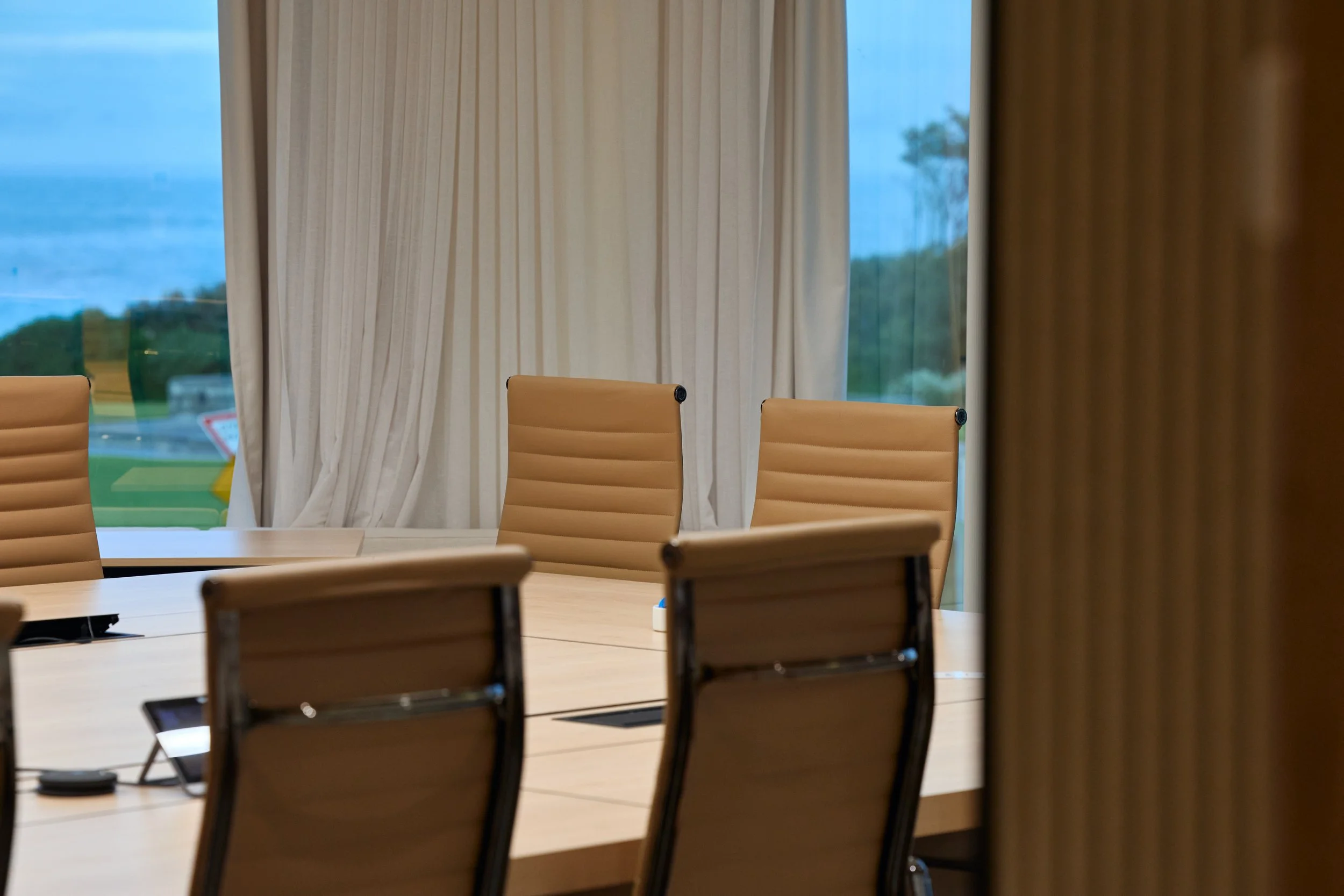 Conference room with beige chairs around a table, large window with floor-length curtains revealing an outdoor landscape.