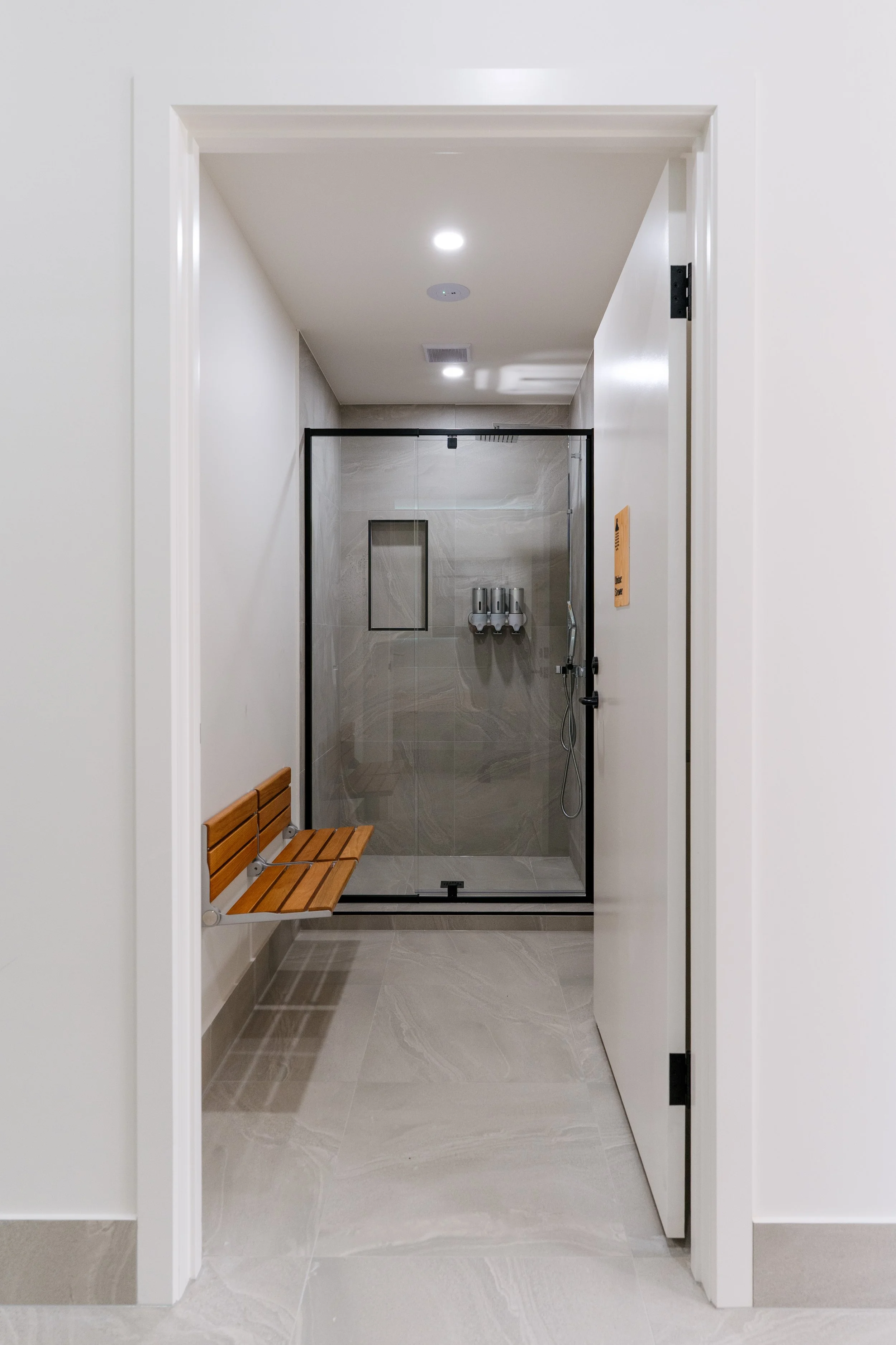 Modern bathroom with gray tiled walls and floor, a glass shower enclosure with a black frame, a wooden bench, and white ceiling with recessed lighting.