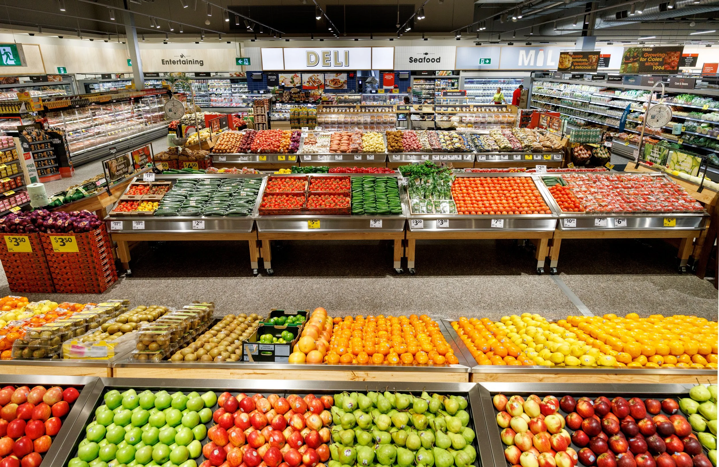 Inside a grocery store with sections labeled Deli, Seafood, and Milling. The store has display shelves with fresh apples, tomatoes, and other produce in a well-lit, organized space.