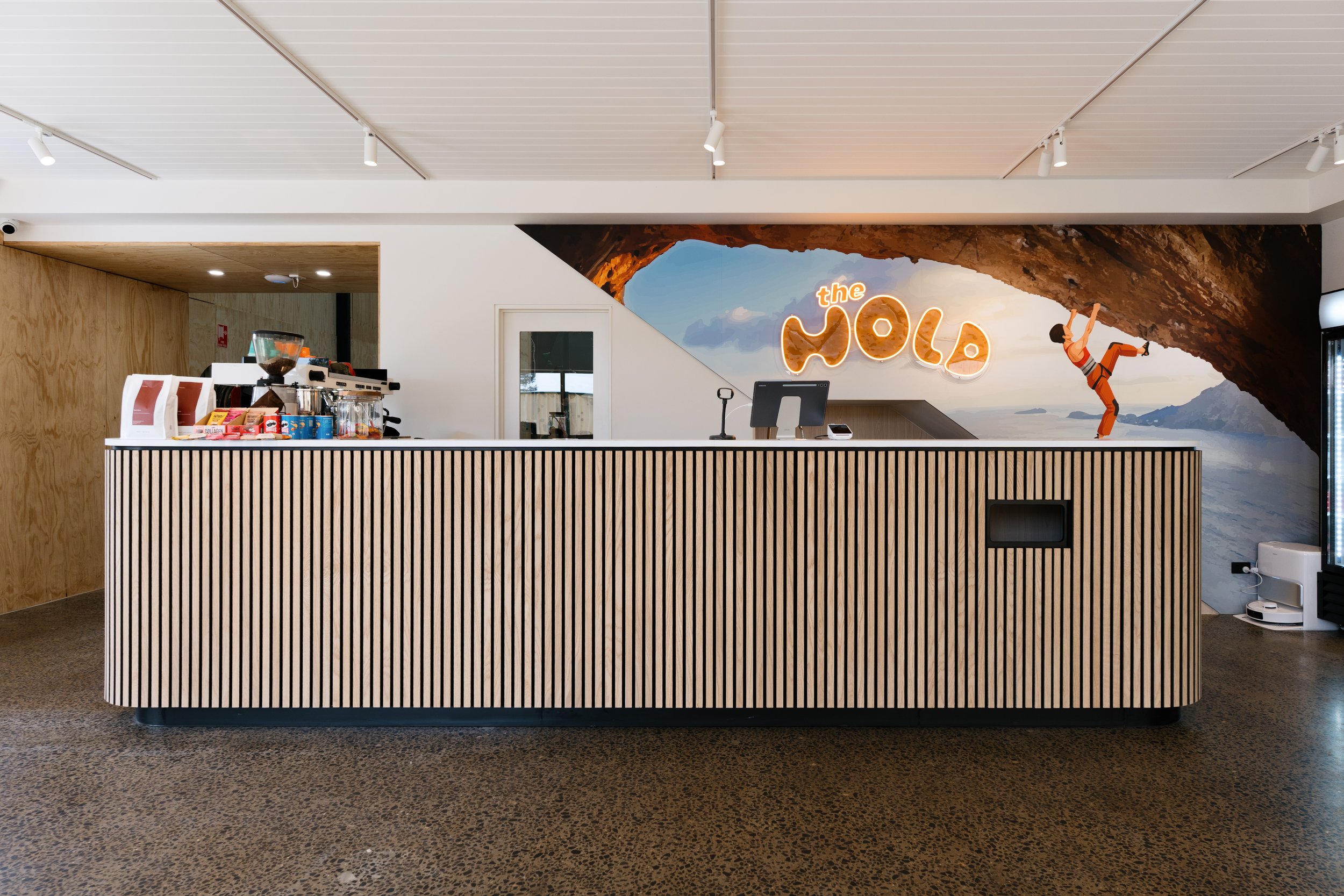 Interior of a modern reception area with a wooden slat reception desk, a coffee station, and a colorful mural wall featuring the words 'the WOLA' and an illustration of a person rock climbing on a mountain over a scenic sky and landscape background.