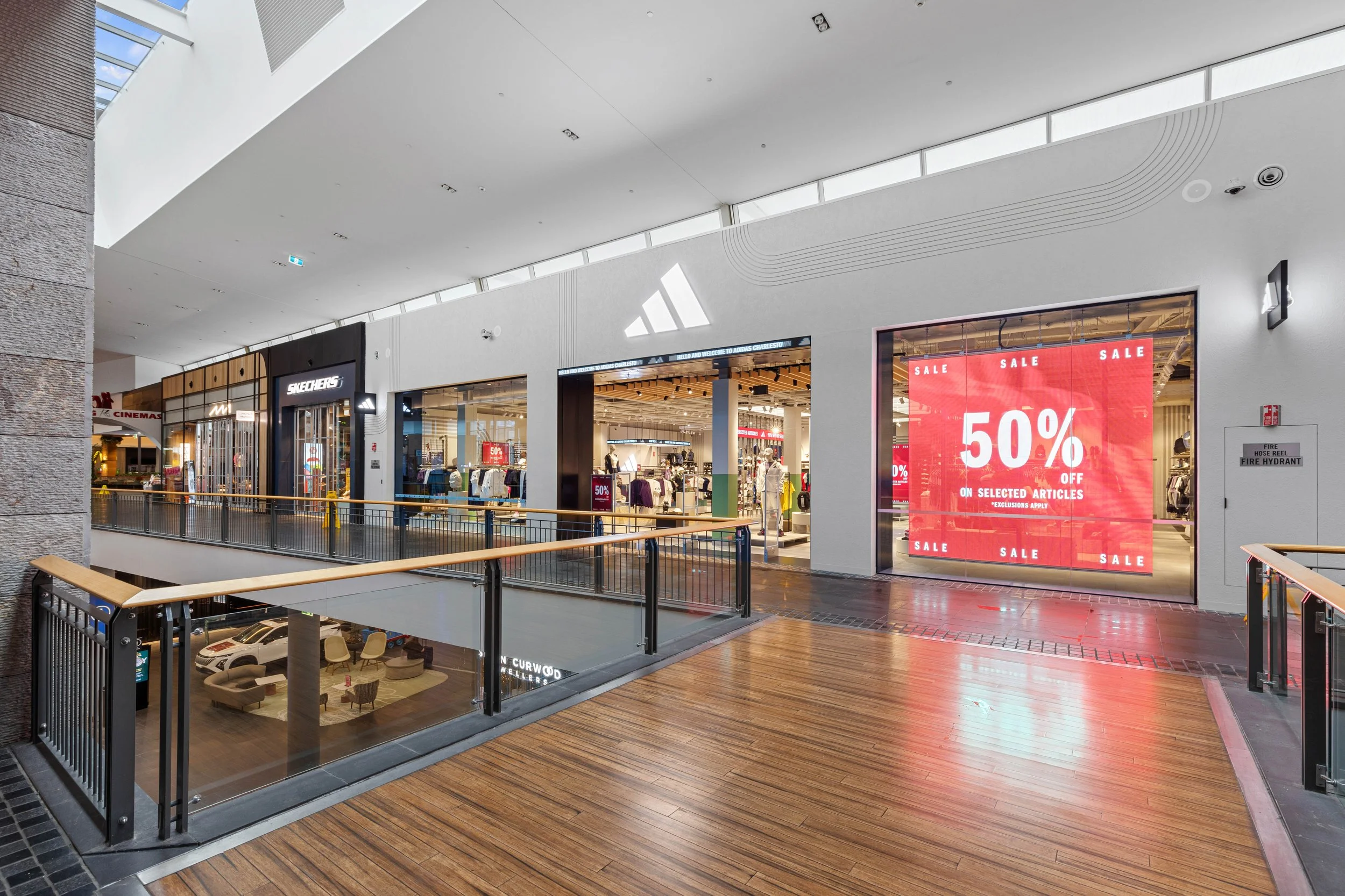 Interior view of a shopping mall with stores and sale signs, including Skechers, with a wooden floor and railings overlooking an underground parking area.