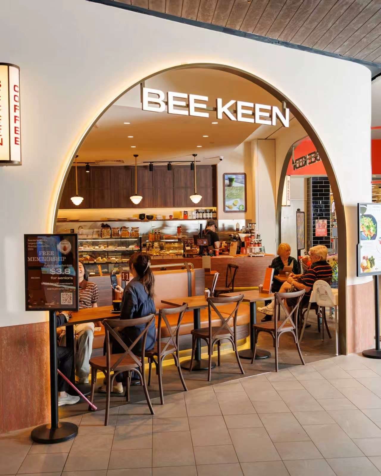 Interior of a Bee Keen cafe with a wooden counter, customers seated at tables and a menu display.