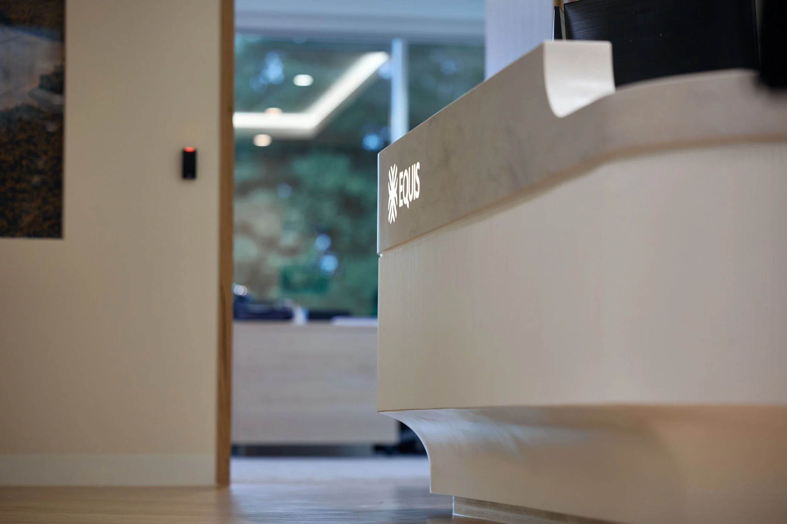 Close-up of a reception desk with a sign on the front, in a modern office or reception area with a window in the background showing trees.