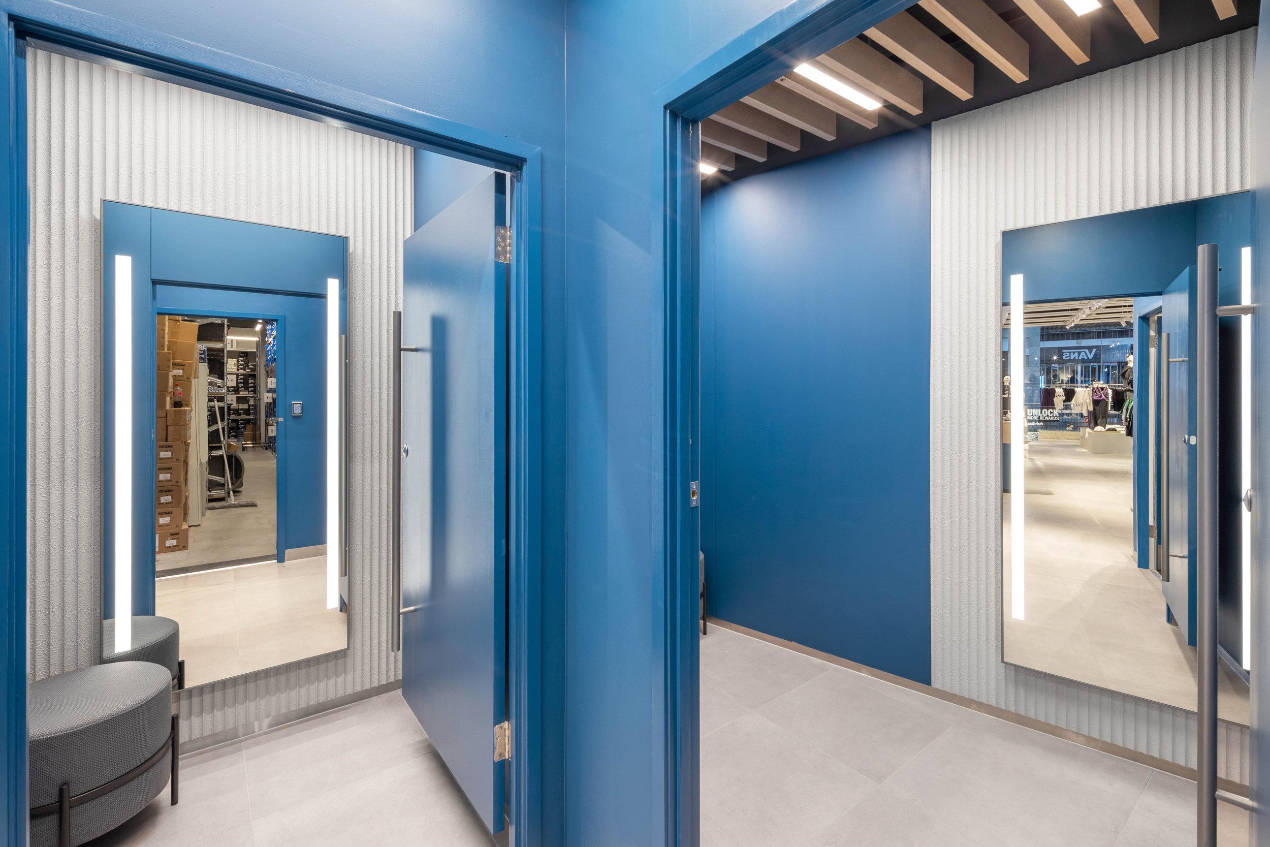 A modern fitting room in a clothing store with blue walls, mirrors, and doors, and a display of shoes and clothing visible in the background.