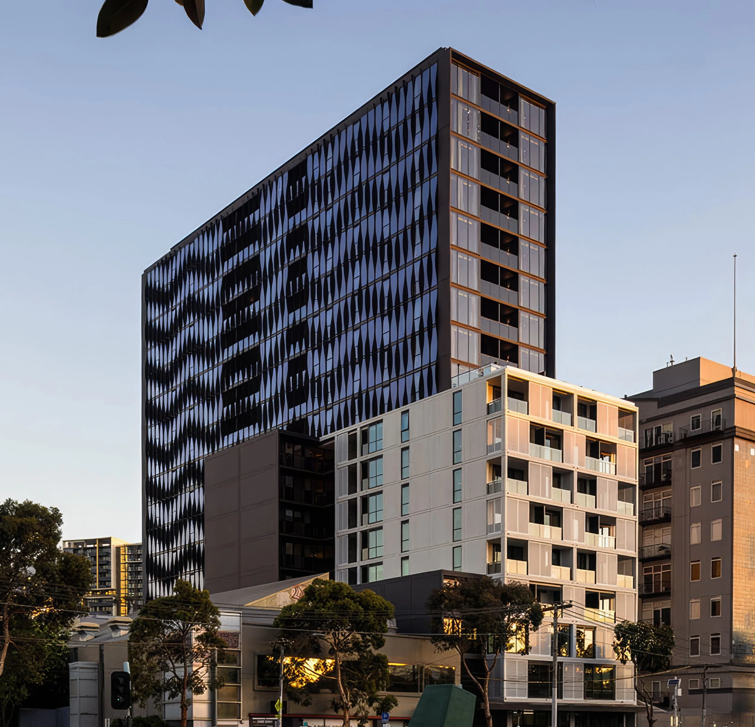 A modern high-rise building with a unique dark facade featuring the shape of slanted diamond patterns. The building has large glass windows and a vertical corner with exposed balcony spaces. The sky is clear and blue, and the lower part of the image shows trees and street-level elements.