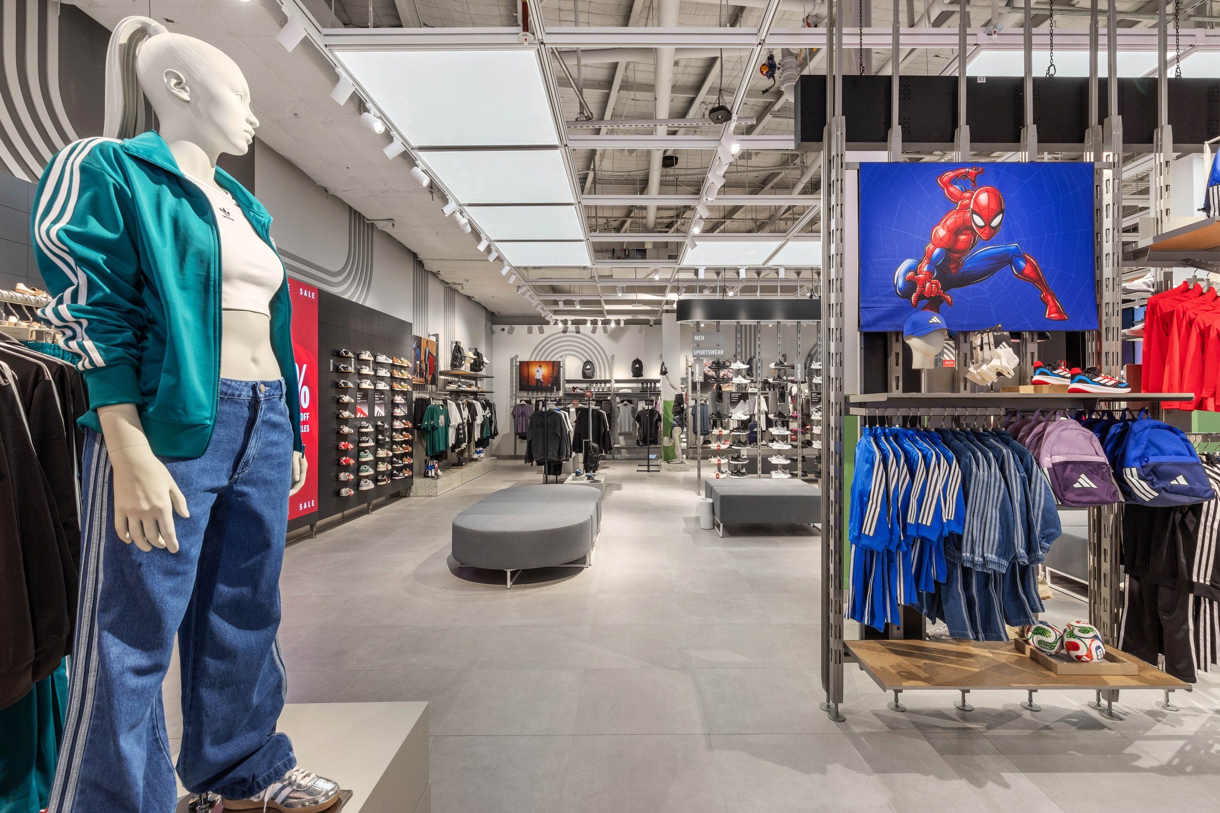 A mannequin wearing a blue Adidas jacket, white shirt, and jeans in a retail store. The store has shelves with shoes, clothes, and backpacks and a large Spiderman poster hanging overhead.