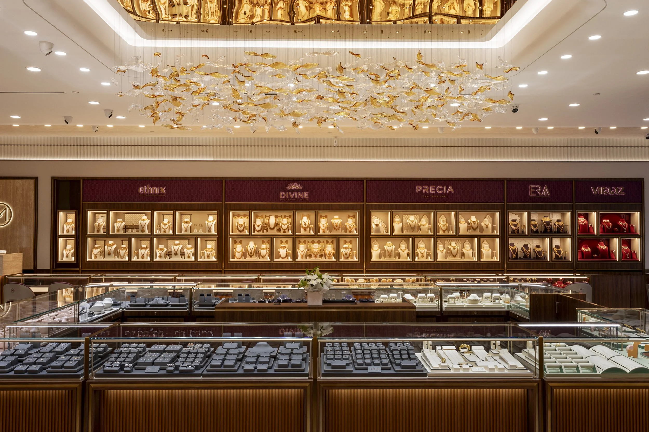 Jewelry store display with necklaces, rings, and earrings showcased in illuminated glass cases against a backdrop of jewelry brand signs and a decorative chandelier overhead.